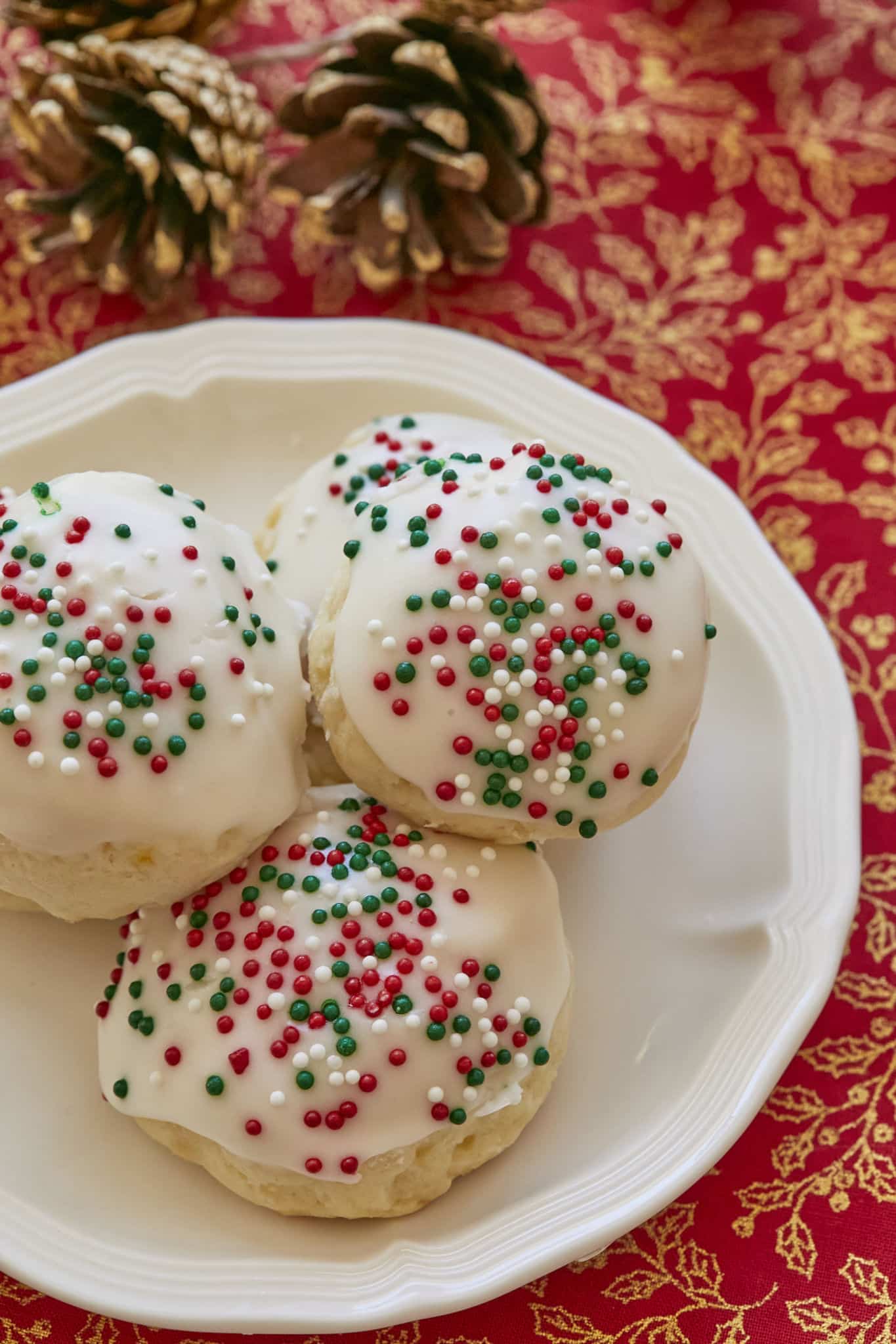 A close-up image of homemade Italian Ricotta Cookies shows a pale cookie topped with vanilla glaze, white, red, and green sprinkles.