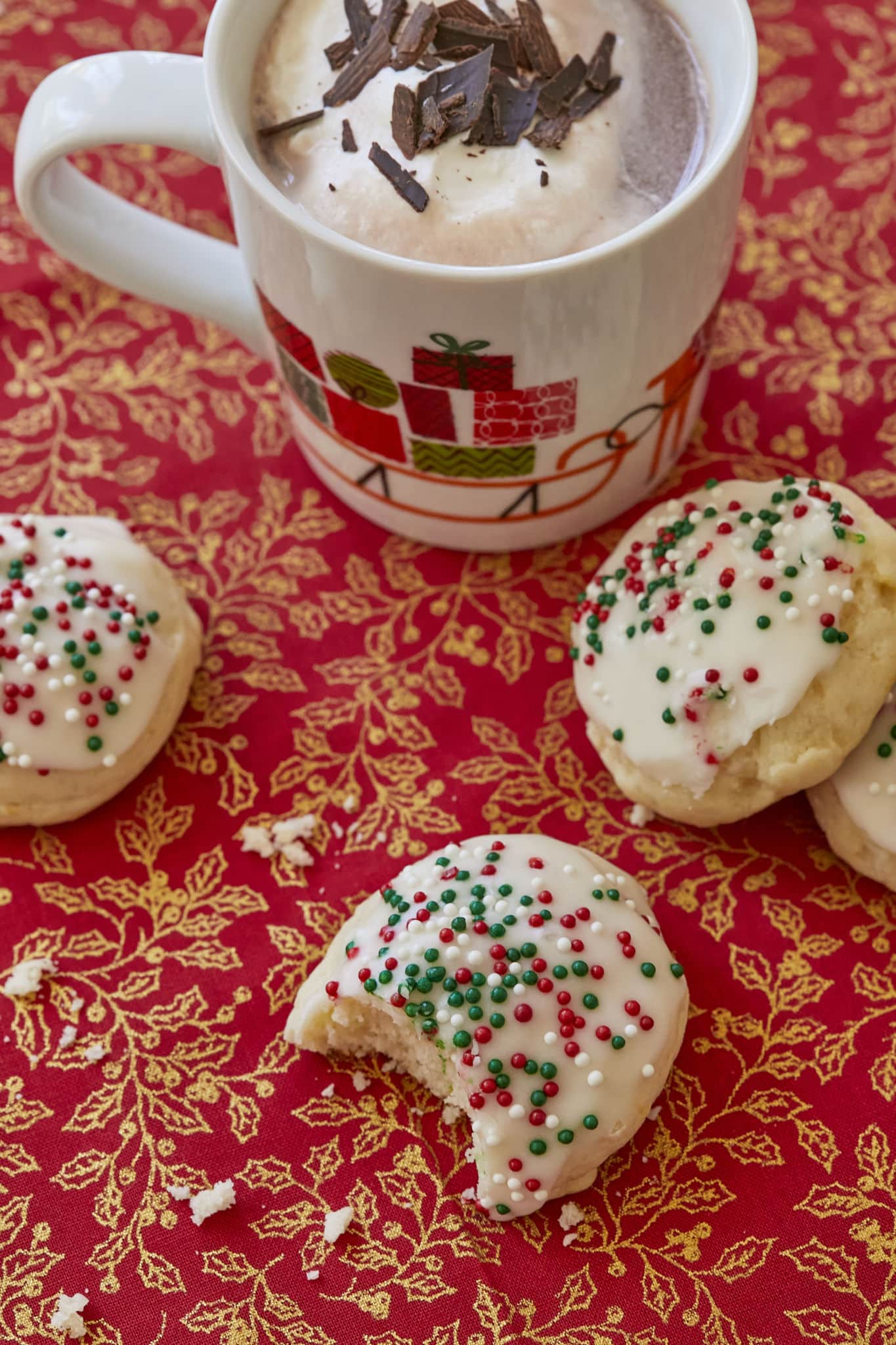 Four Italian Ricotta Cookies, one with a bite taken out of it showing its cake-like texture, are on a red table cloth with golden accents of holly berries. There is a mug of hot chocolate above them, topped with homemade whipped cream and chocolate shavings.
