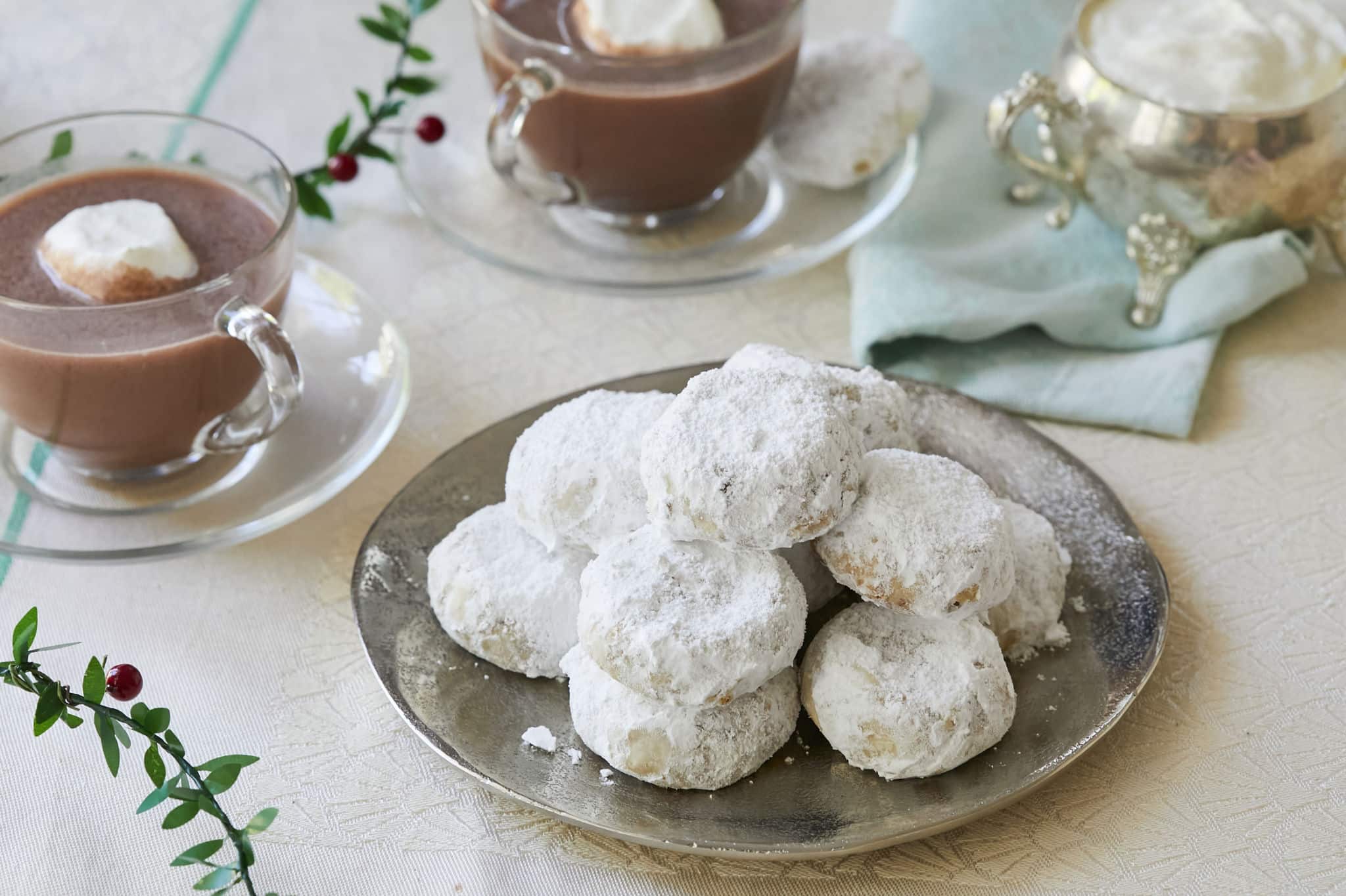 Homemade Greek Kourabiedes are presented on a silver plate next to two clear mugs of hot chocolate topped with marshmallows. The Kourabiedes are coated in powdered sugar.