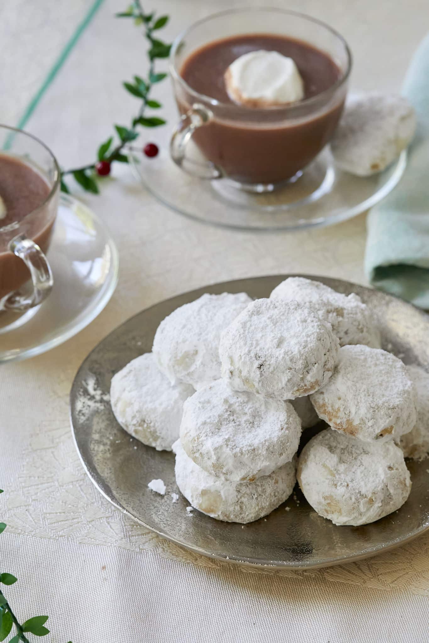Kourabiedes, coated in powdered sugar, are served on a silver plate on top of a table decorated with holly berries. Next to the Christmas cookies are two mugs of hot chocolate. 