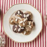 A plate of homemade Lebkuchen cookies are presented on a red and white striped table cloth next to three mugs of hot chocolate. Some of the Lebkuchen cookies are covered in a glaze while others are covered in a chocolate glaze. They are all topped with slivered almonds.