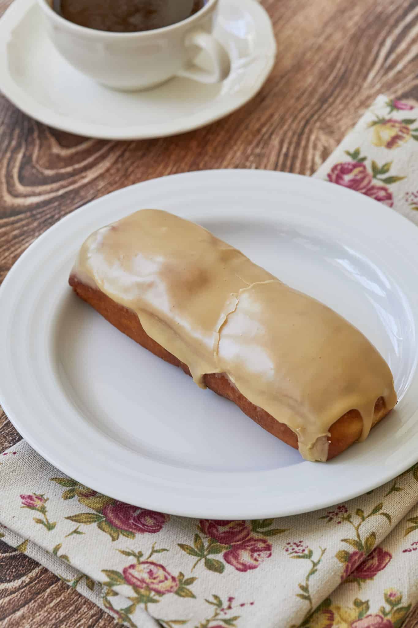 A single maple bar donut is served on a white plate near a black cup of coffee.