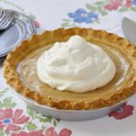 A homemade Maple Cream Pie is served on a floral tablecloth. The homemade pie has a big dollop of fresh whipped cream piled on top.
