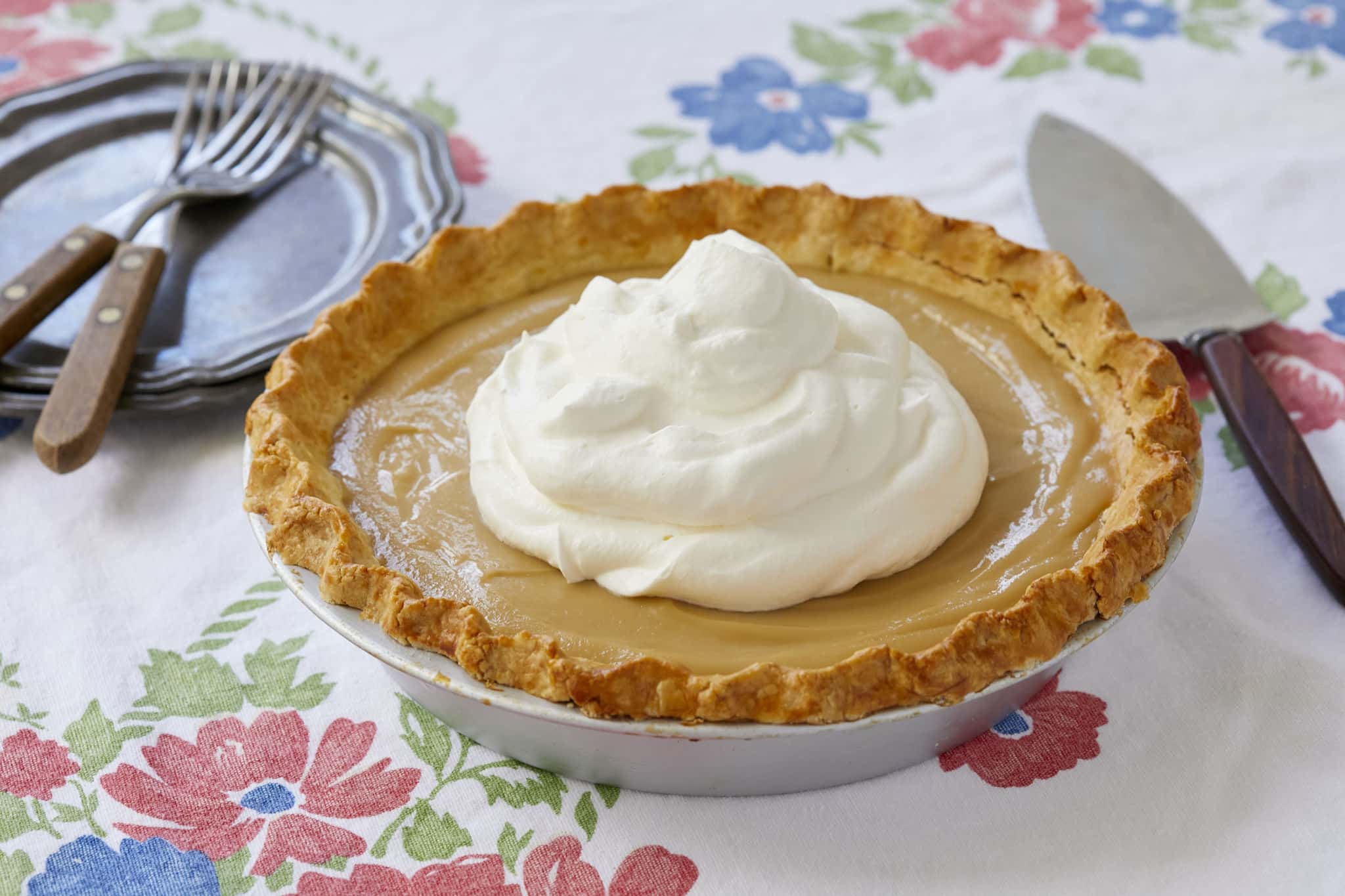 A homemade Maple Cream Pie is served on a floral tablecloth. The homemade pie has a big dollop of fresh whipped cream piled on top.