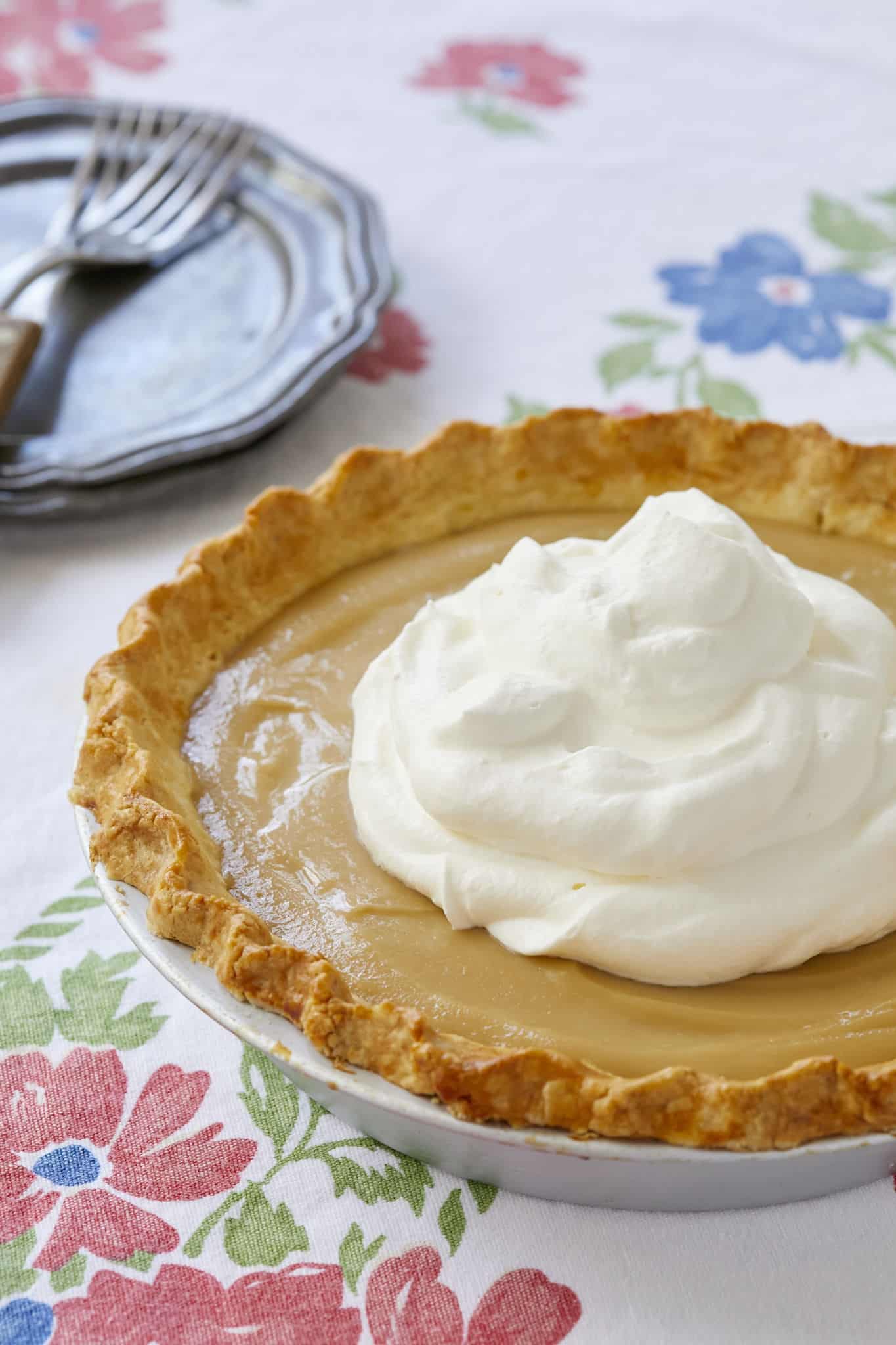 Maple Cream Pie is displayed on a floral tablecloth under a large dollop of homemade whipped cream. 