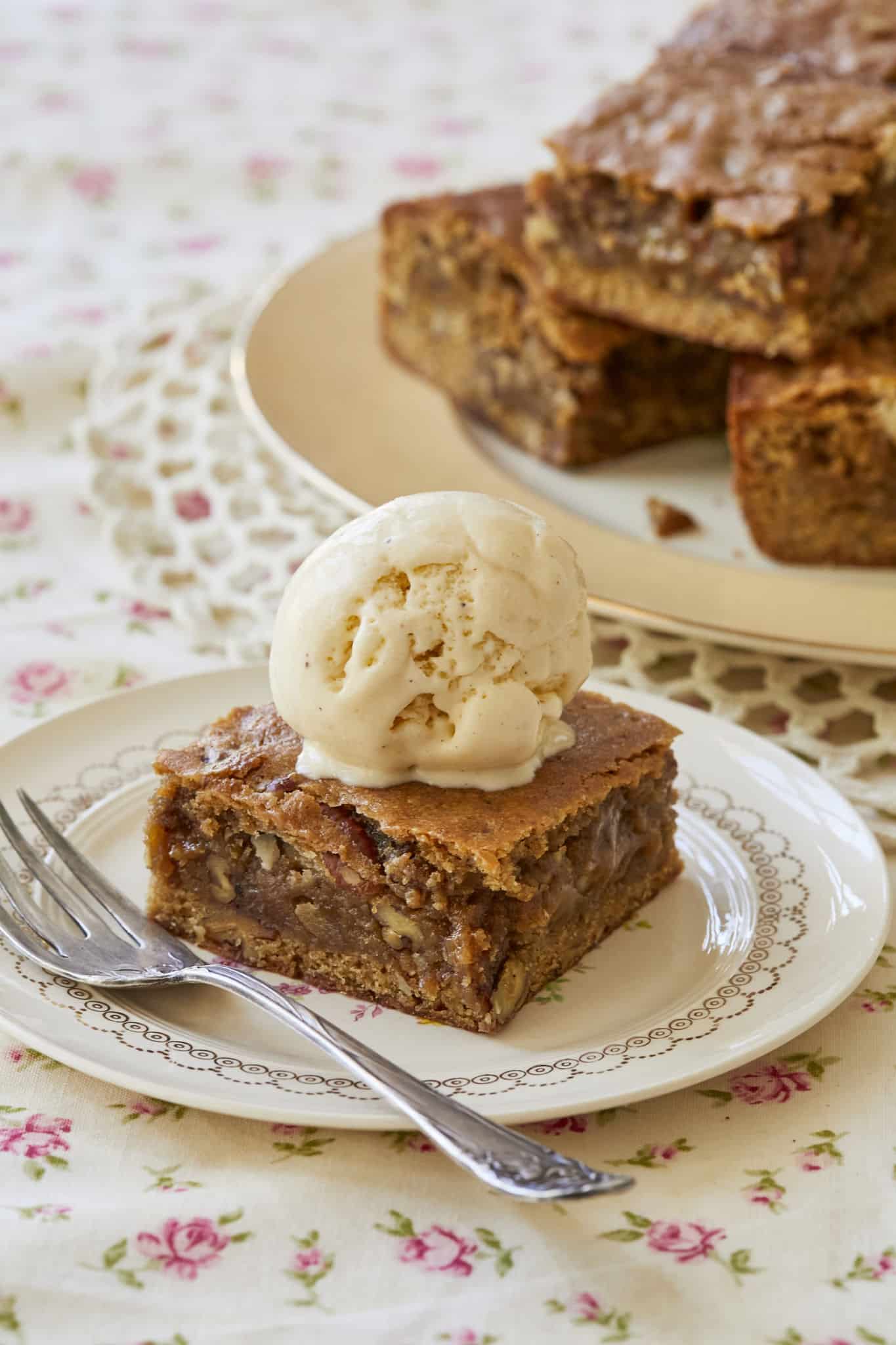 Homemade maple pecan blondies are stacked in the background of a photo of one served on fine china, topped with vanilla ice cream.