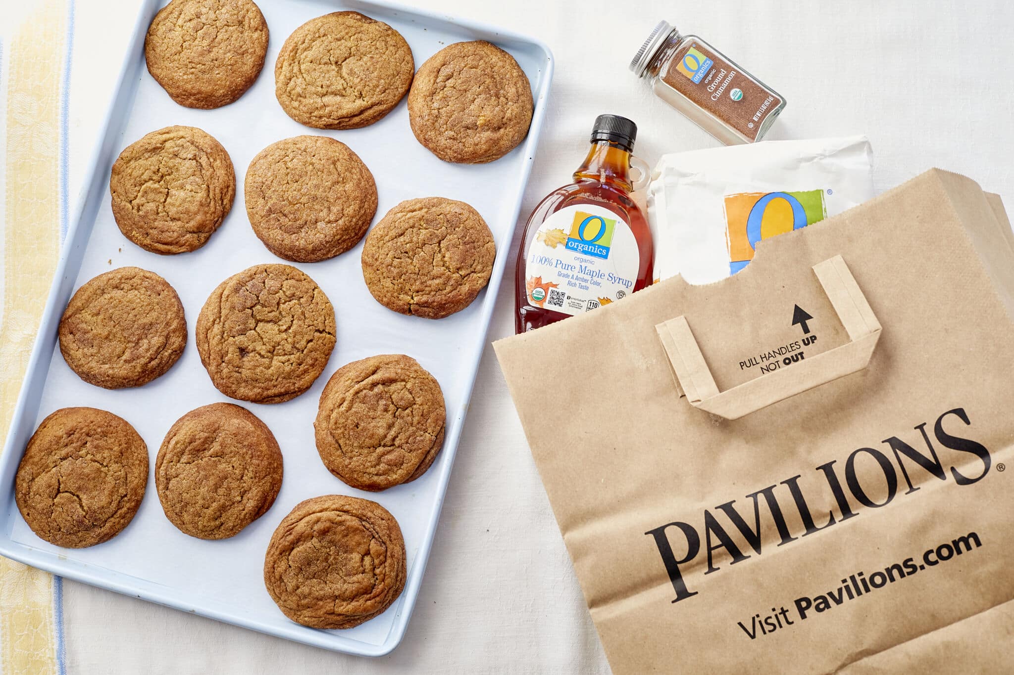 A tray of maple pumpkin cookies on a baking sheet next to a brown paper shopping bag with ingredients spilling out.