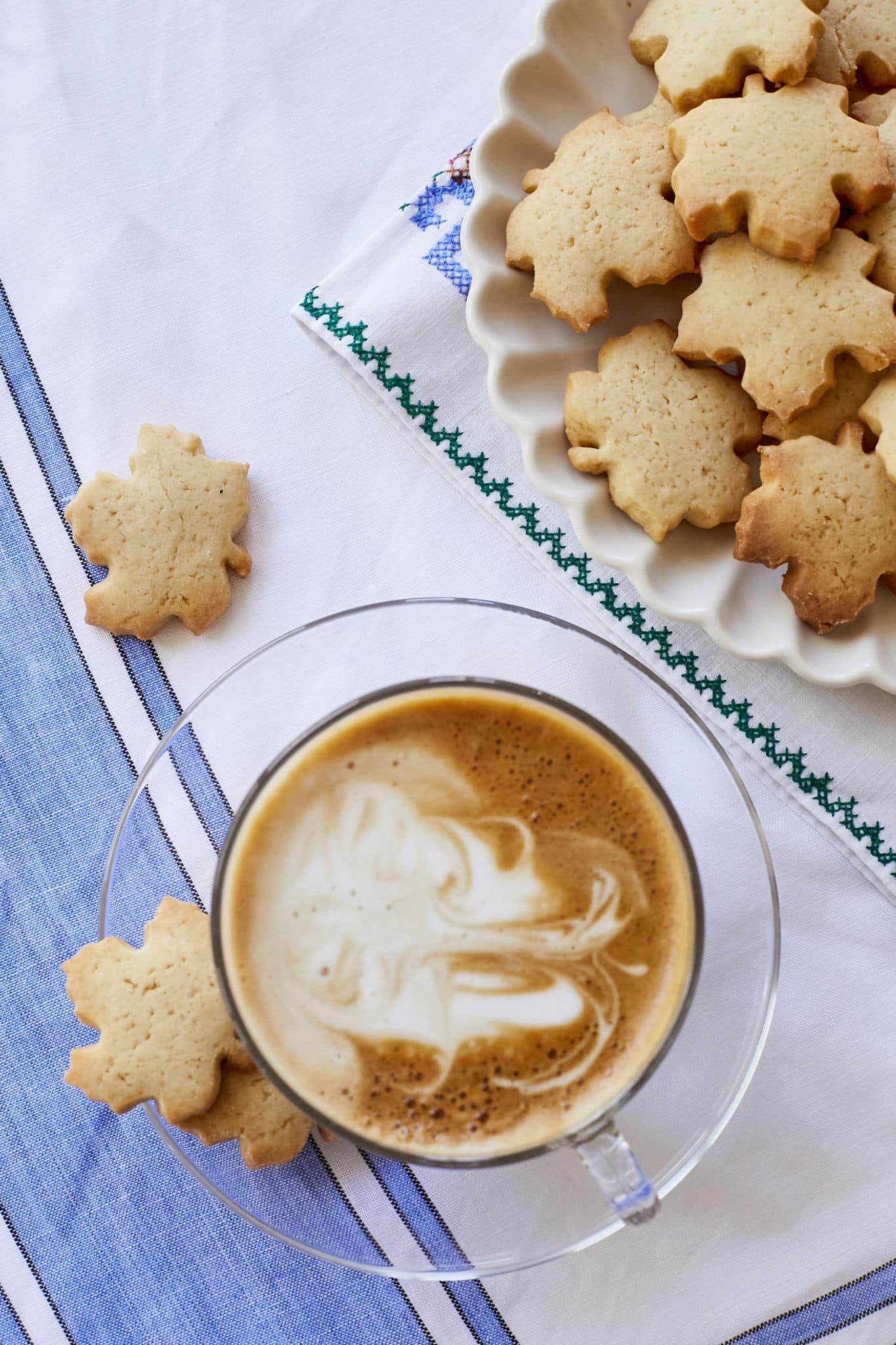 A plate of crunch maple cookies on a white plate next to a latte with individual maple cookies strewn about the table.