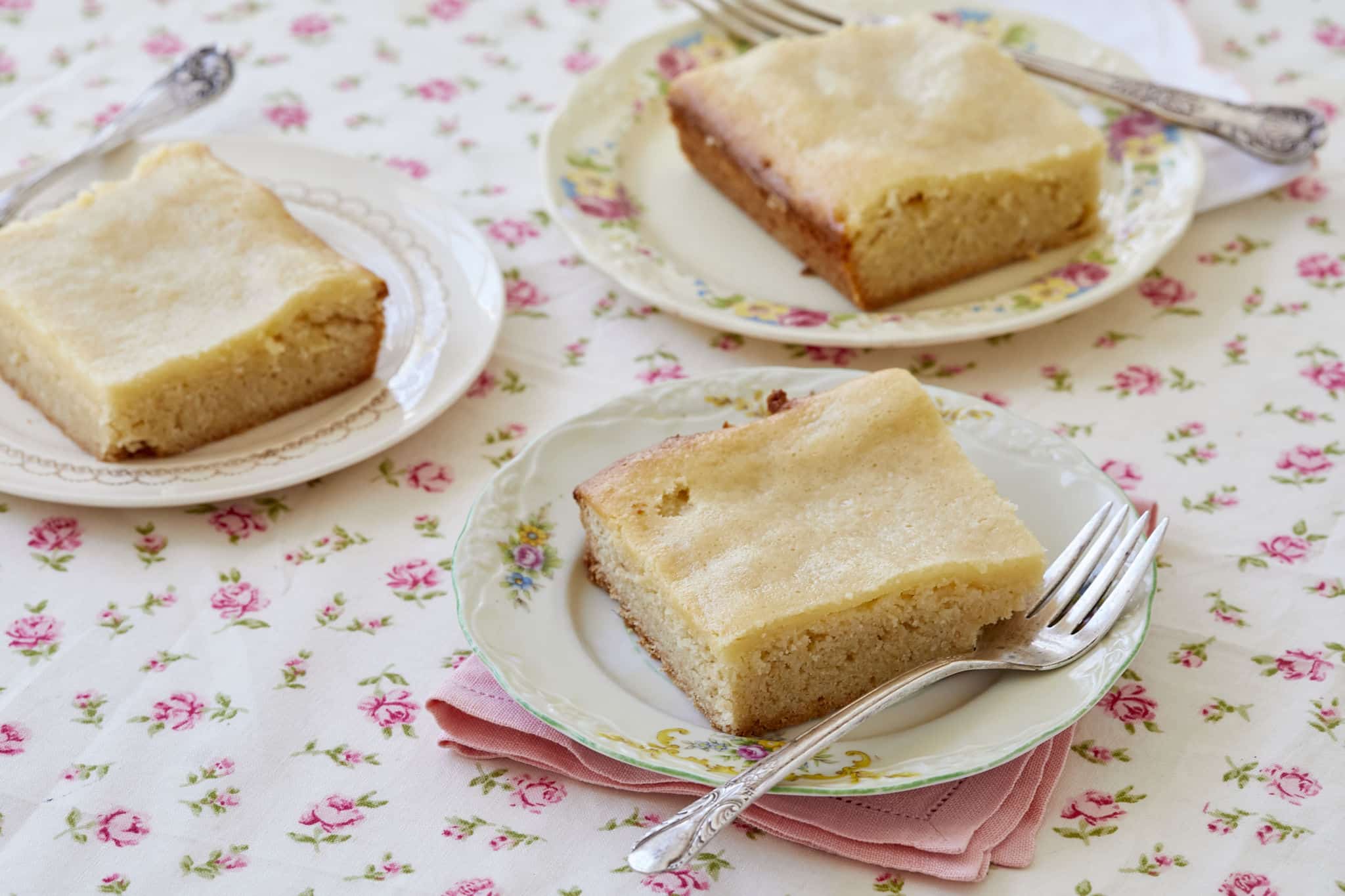 Three pieces of maple gooey butter cake on individual plates with forks, on a floral tablecloth.