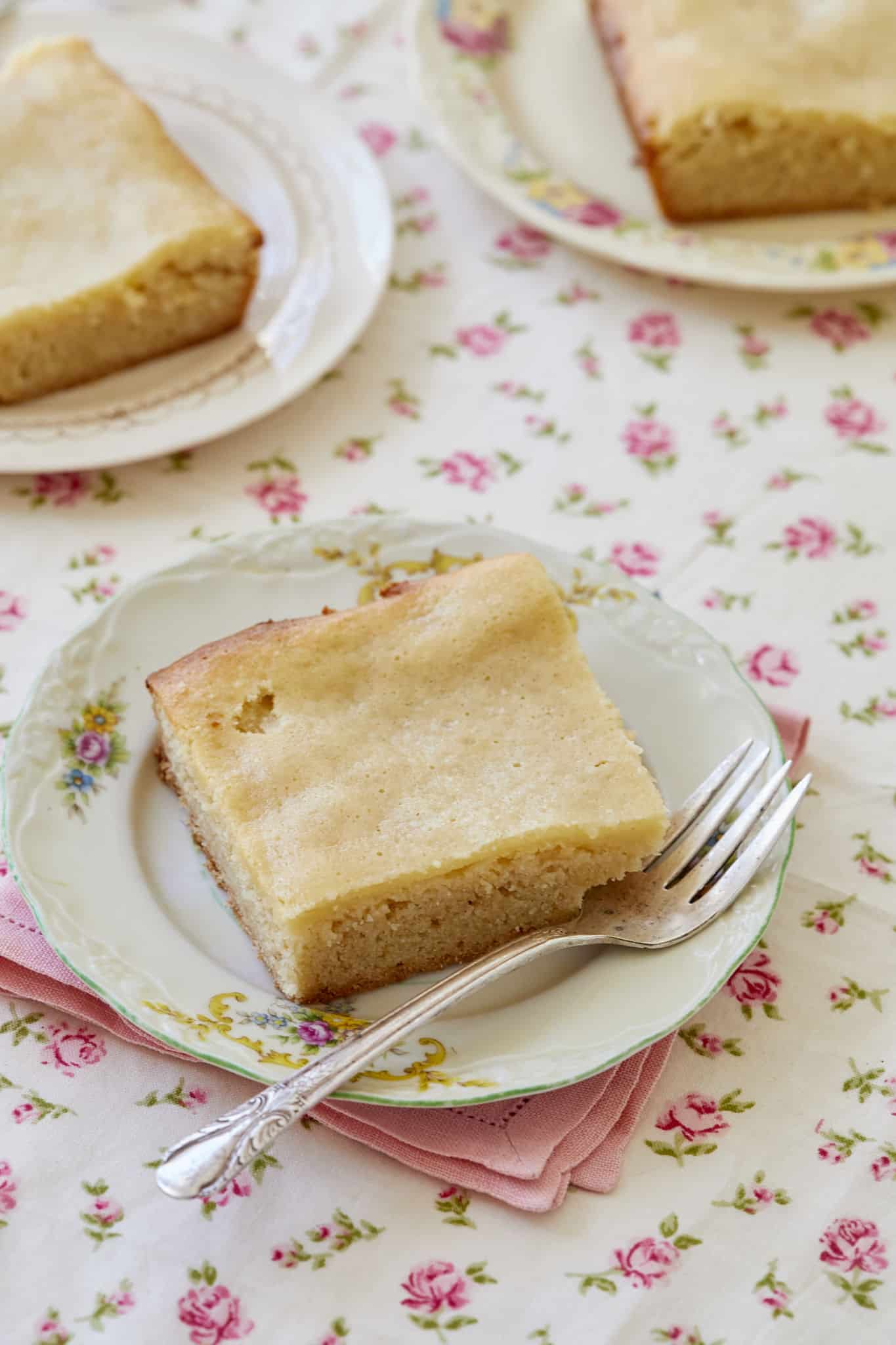 Three pieces of maple gooey butter cake on individual white plates with a fork, on top of pink napkins and a floral tablecloth.