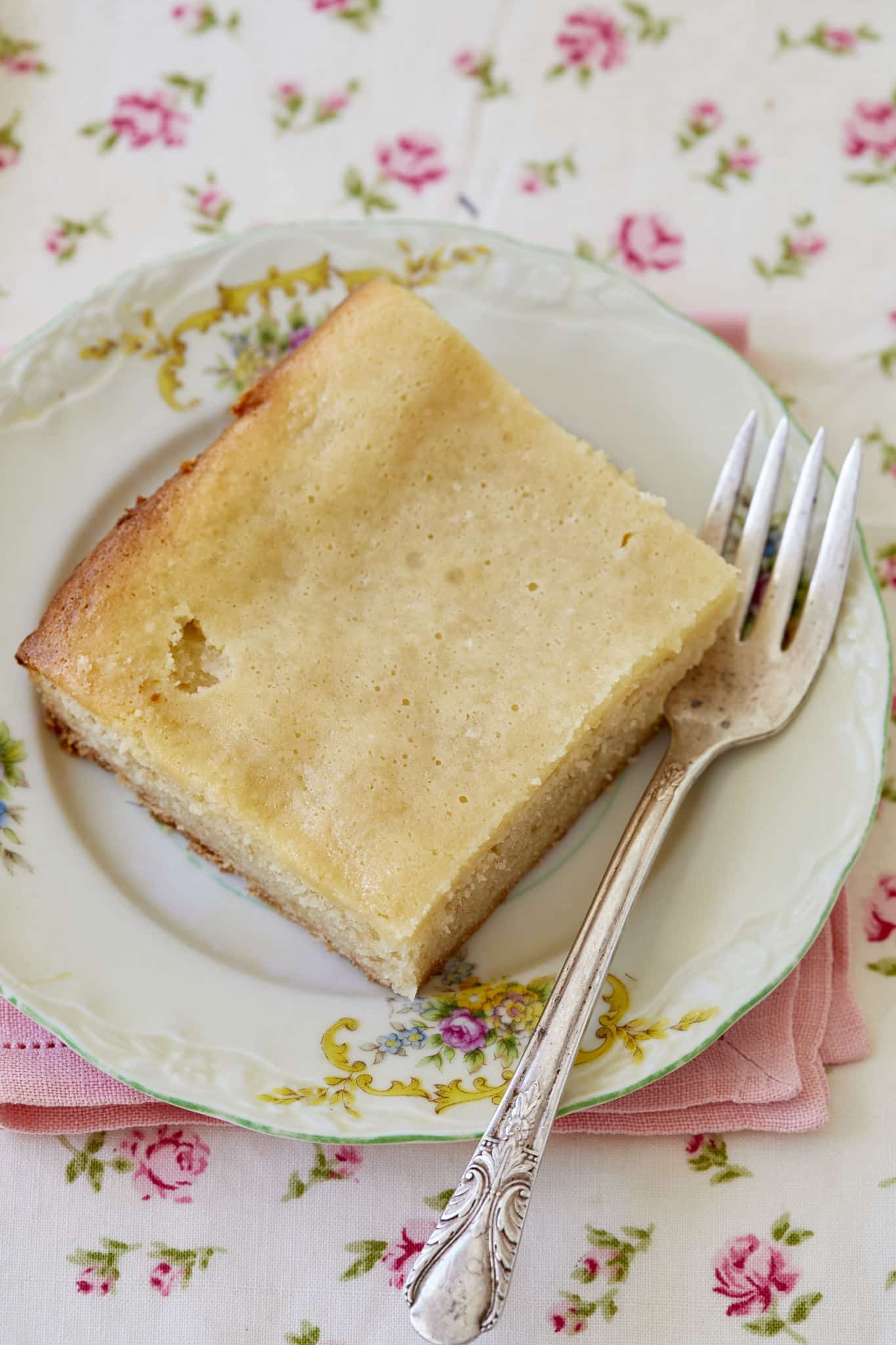 A close-up overhead shot of one piece of maple gooey butter cake on a vintage plate with a fork, on a pink napkin and floral tablecloth.