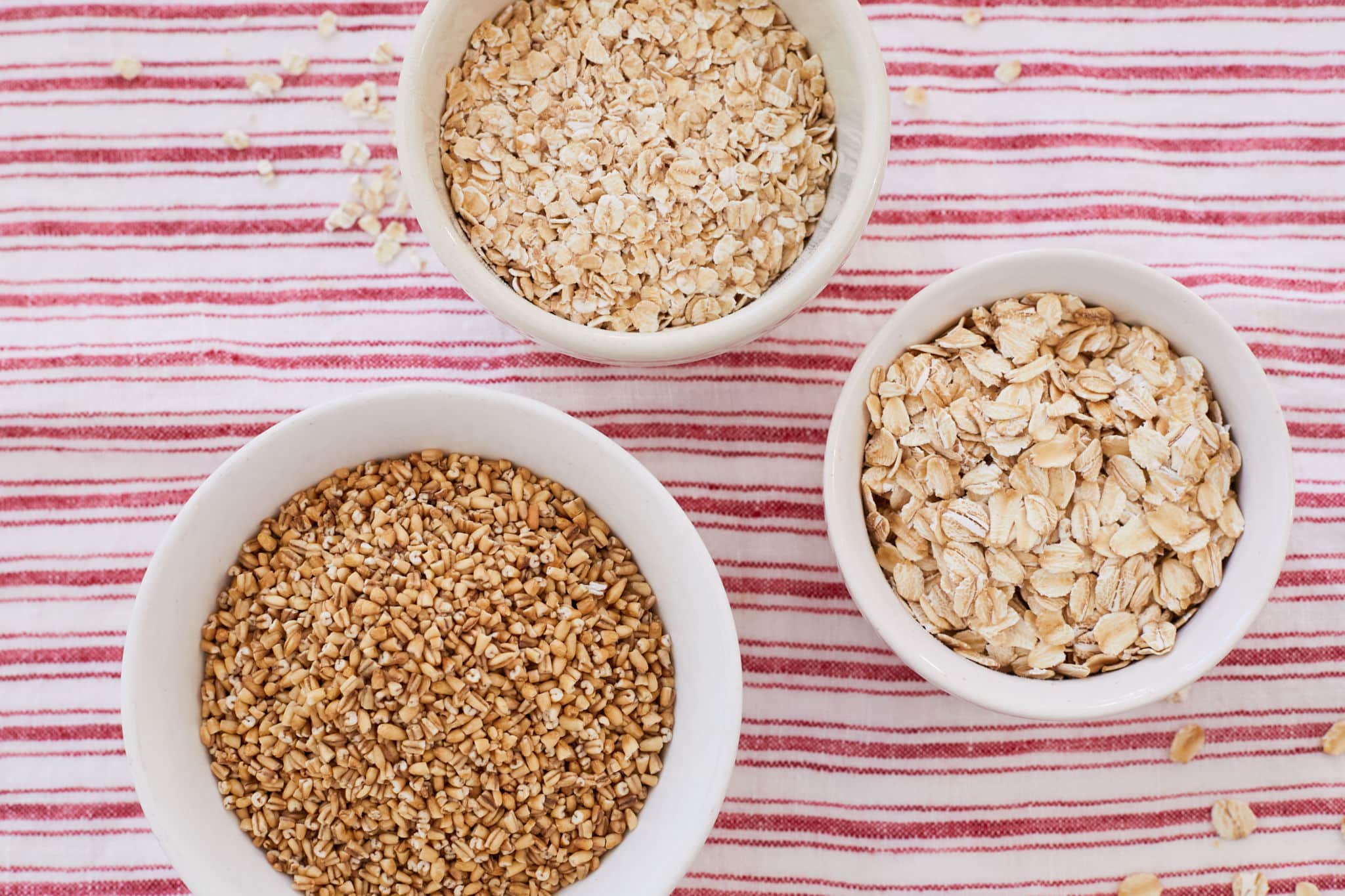 Three different types of oats are displayed in white bowls. At the top is the finest of the three, instant oats, to its right, in the middle, are old fashioned oats, and to the bottom left are oat grains.