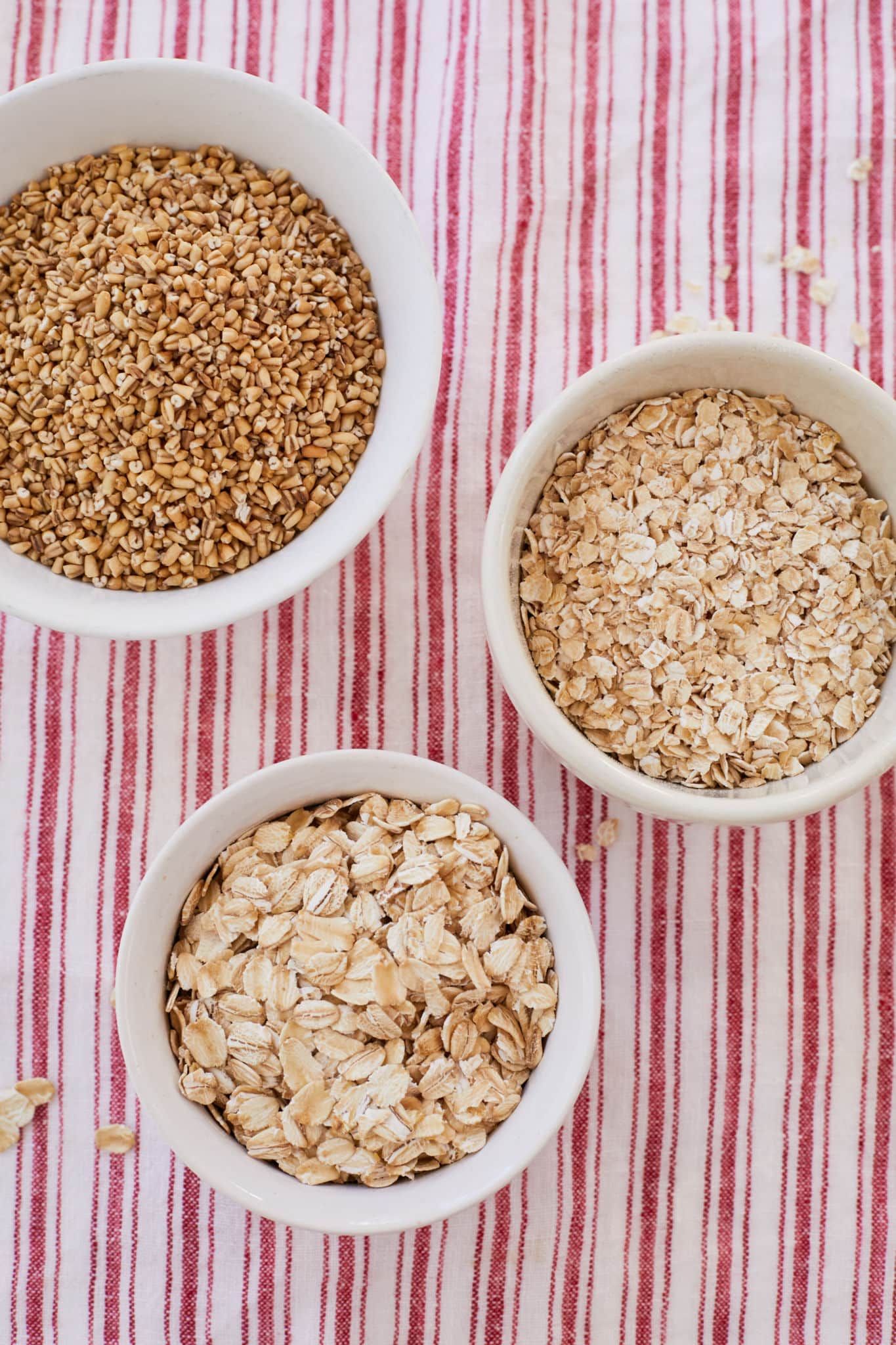 Three different types of oats are displayed in white bowls. On the top left is whole oat bran, toward the middle on the right side are instant oats, and at the bottom toward the left are old-fashioned oats. 