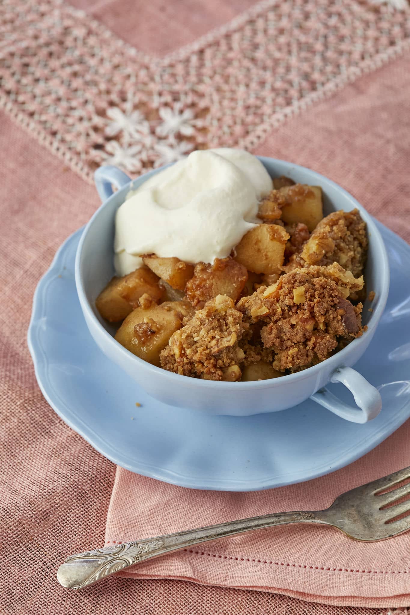 Homemade pear crumble is served in a blue cup on top of a blue saucer with a dollop of whipped cream.