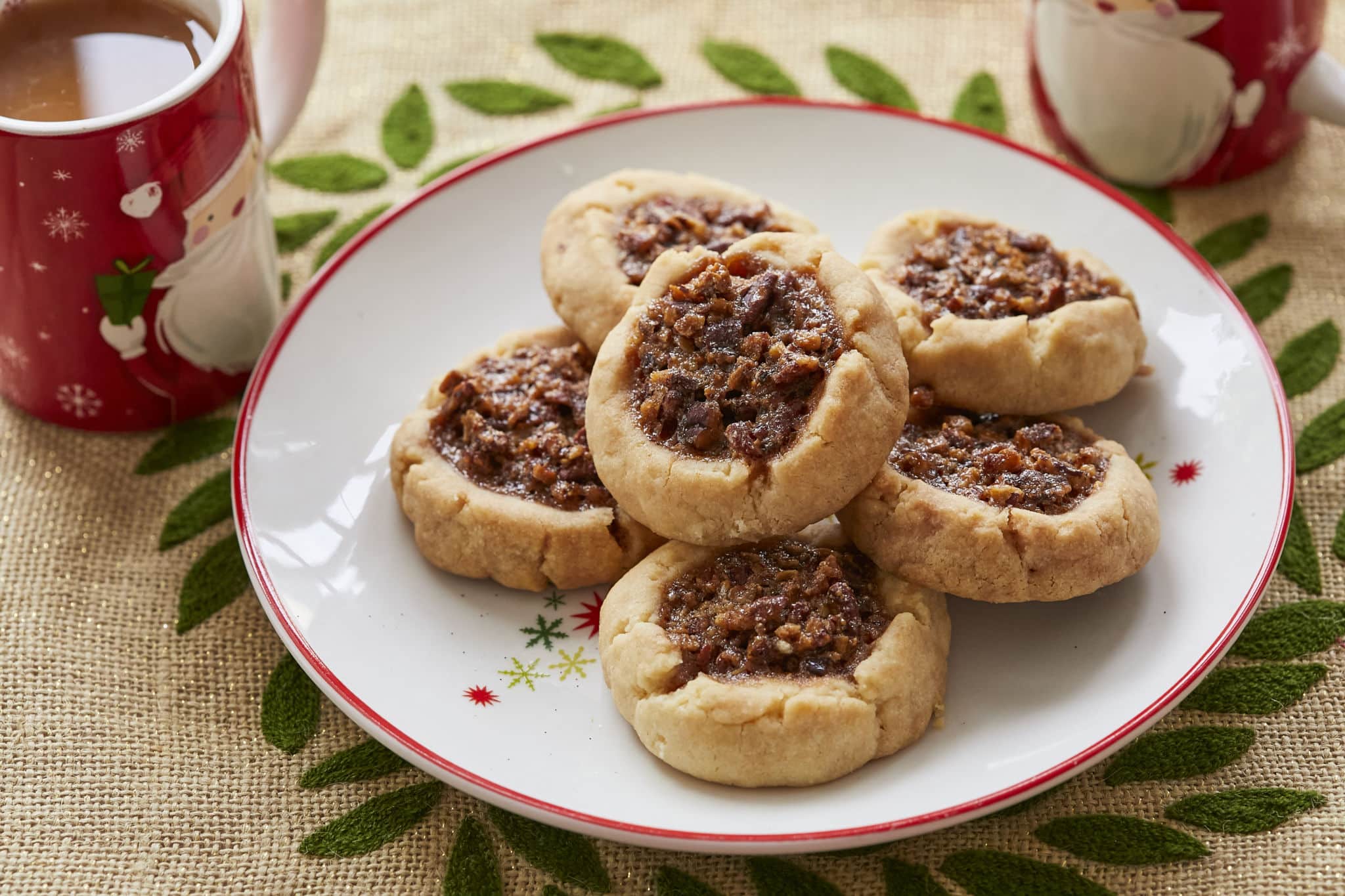 Homemade Pecan Pie Thumbprint Cookies are served on a white dish next to a mug of hot chocolate. The thumbprint cookies are dimpled in the middle and filled with a pecan pie filling.