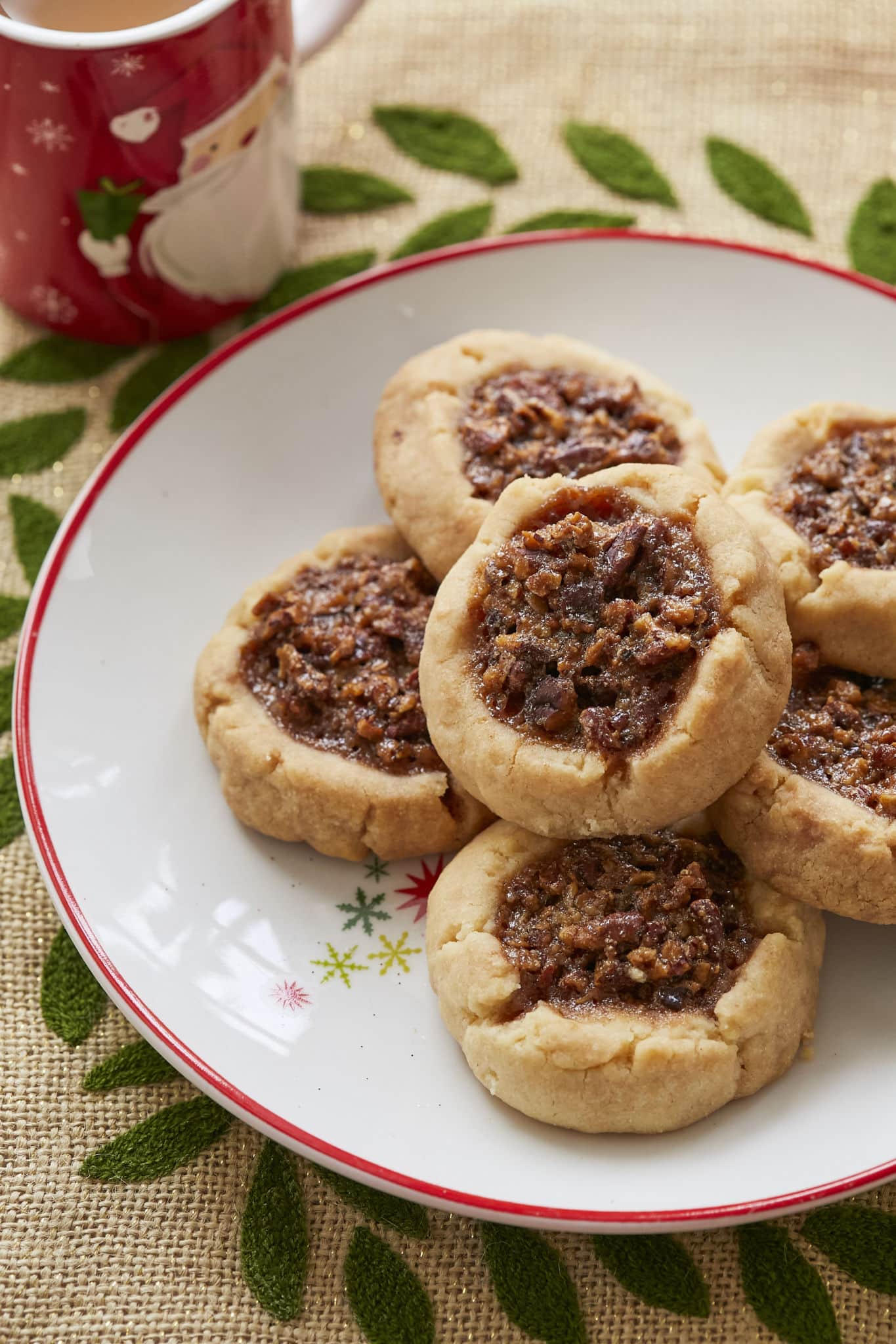 A dish of Pecan Pie Thumbprint Cookies, shortbread cookies filled with pecan pie filling, are served alongside a mug filled with hot chocolate.