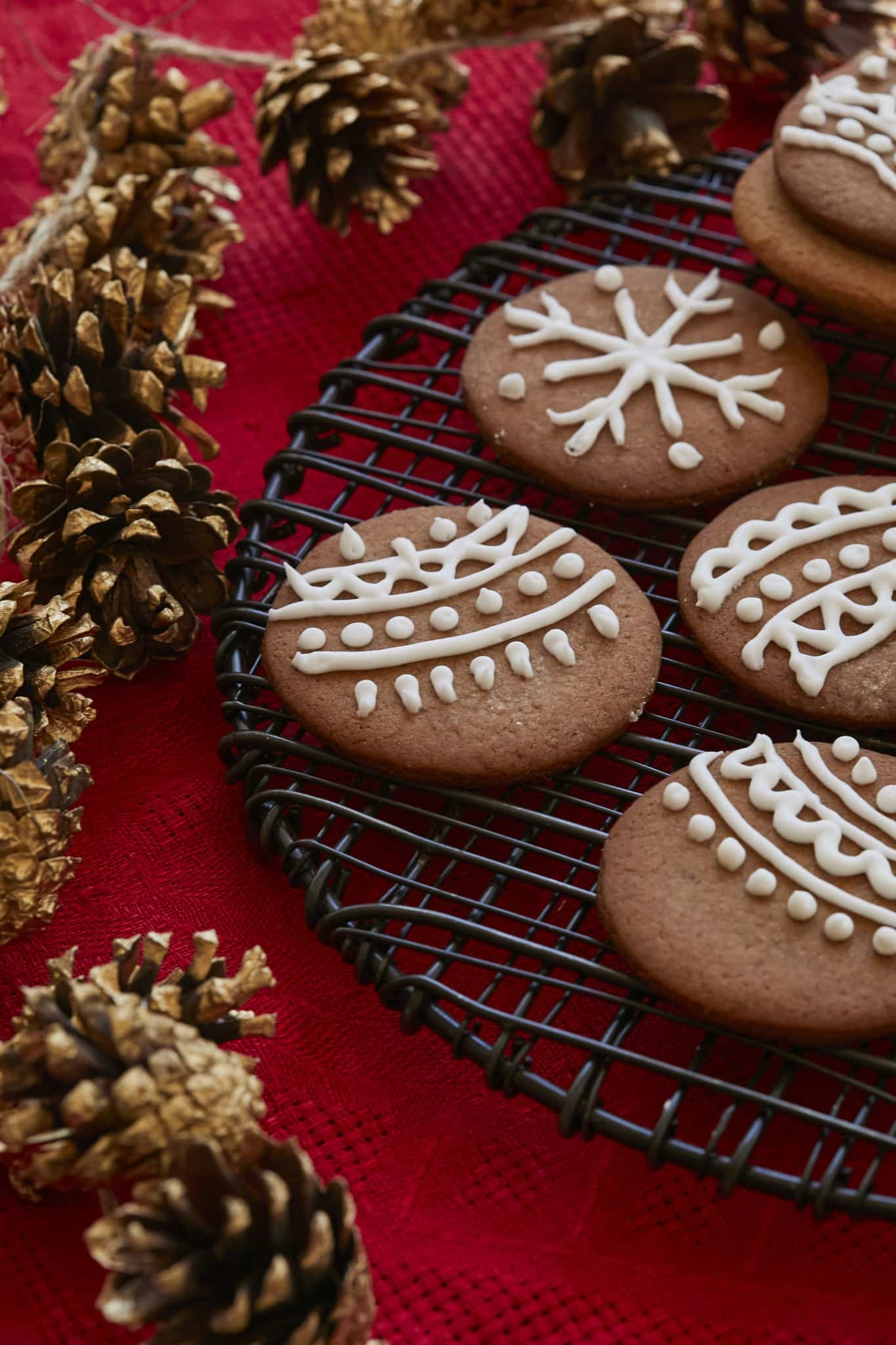 Crispy gingersnaps decorated with white royal icing are shown on a holiday table next to pine cones. 