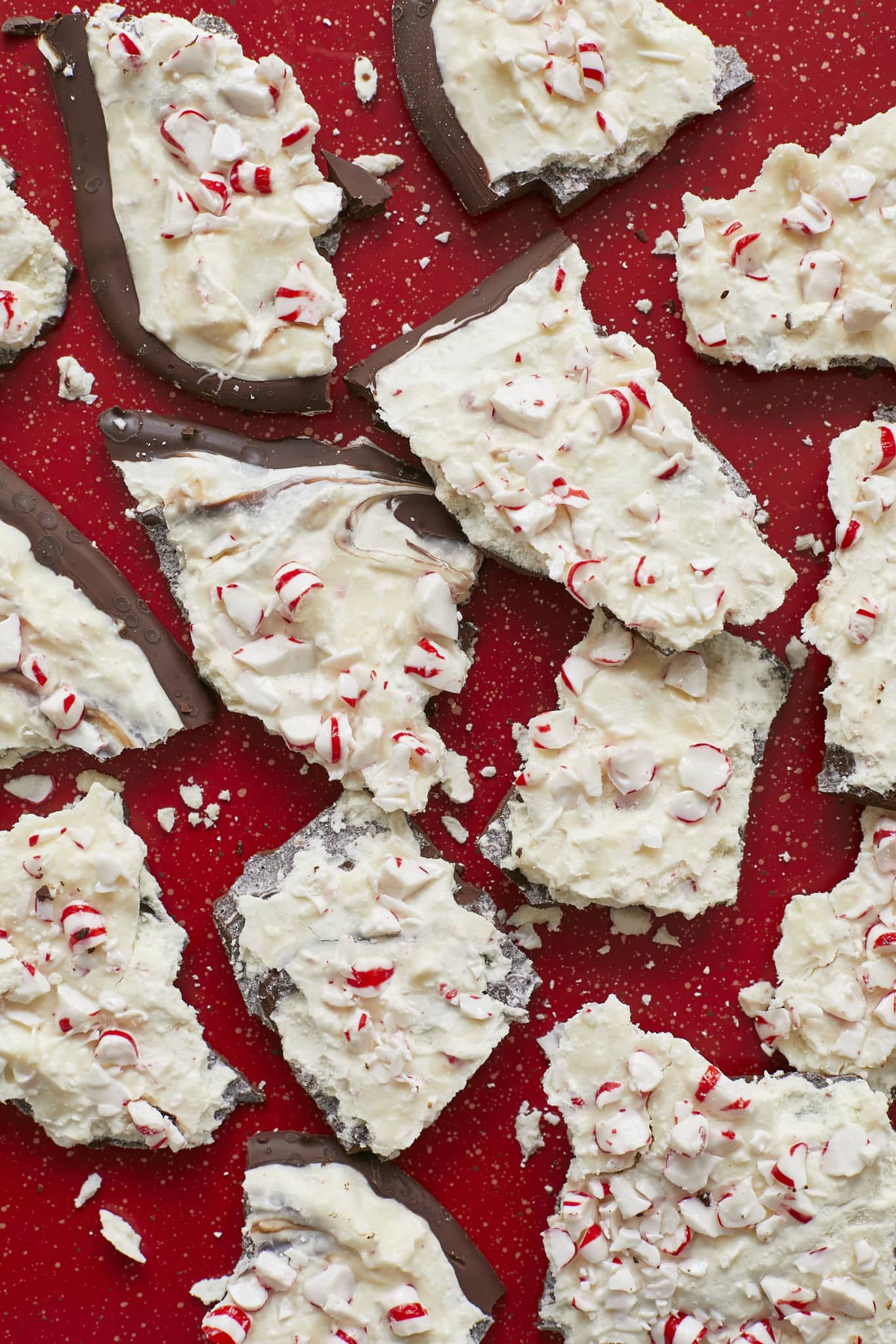 Homemade Peppermint Bark, broken up into pieces, are served on a red table. The bark has layers of dark chocolate and white chocolate and is topped with crushed candy canes.