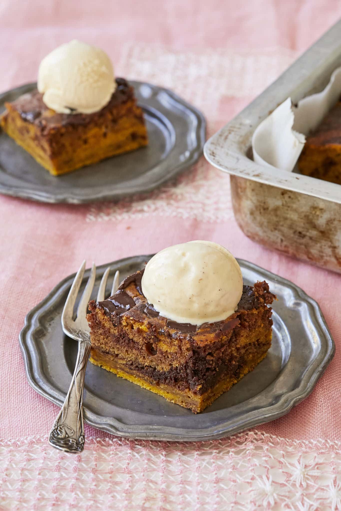 Two pieces of marbled pumpkin brownies on individual silver plates with scoops of vanilla ice cream on top, next to a tray of baked brownies.
