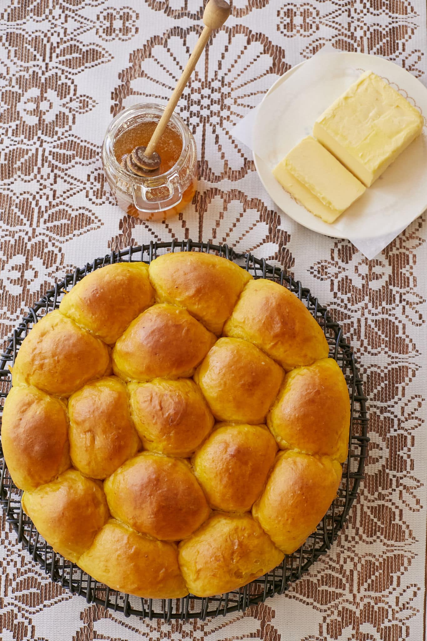 A batch of pumpkin dinner rolls are cooling on a wire rack. Next to the rolls is a plate with butter and a jar of honey. 