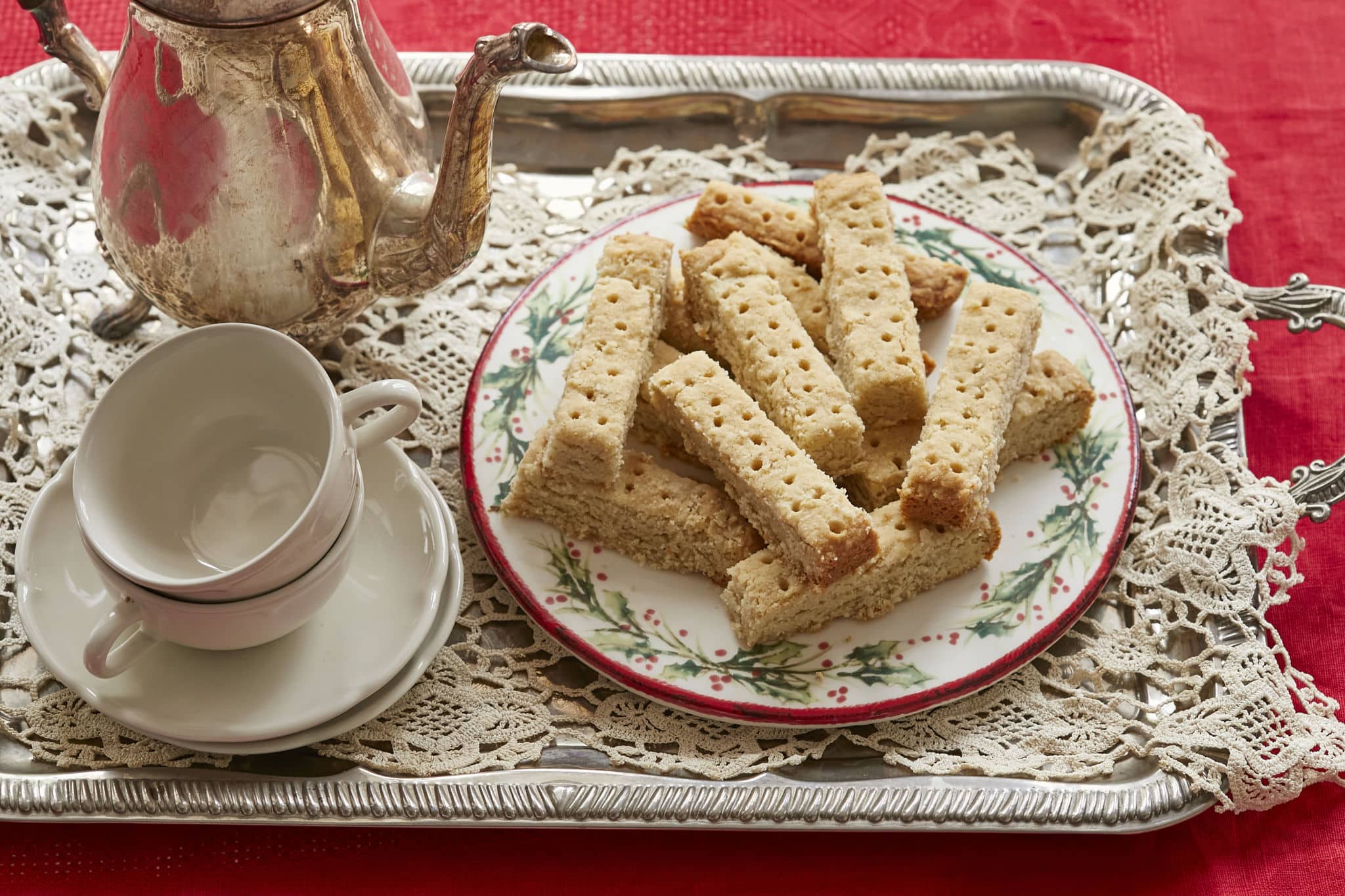 A plate of homemade Scottish Shortbread is presented on a holiday plate, decorated with holly berries, on a plater with an empty tea cup and a silver tea pot