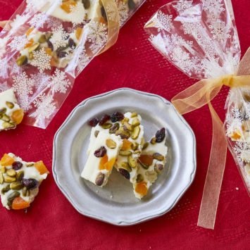 Homemade White Chocolate Bark, topped with dried cranberries, dried apricots, and pistachios, are served on a plate next to festive gift bags filled with pieces of the bark.