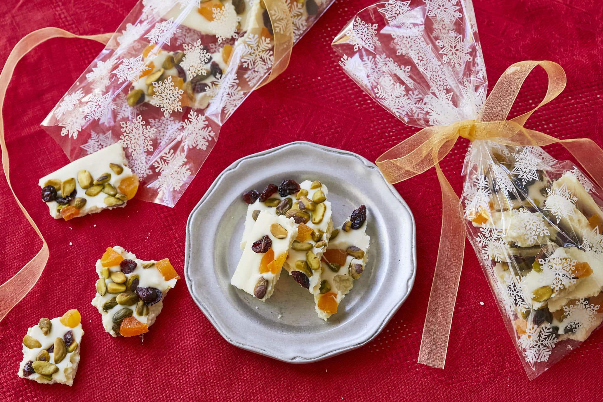 Homemade White Chocolate Bark, topped with dried cranberries, dried apricots, and pistachios, are served on a plate next to festive gift bags filled with pieces of the bark.