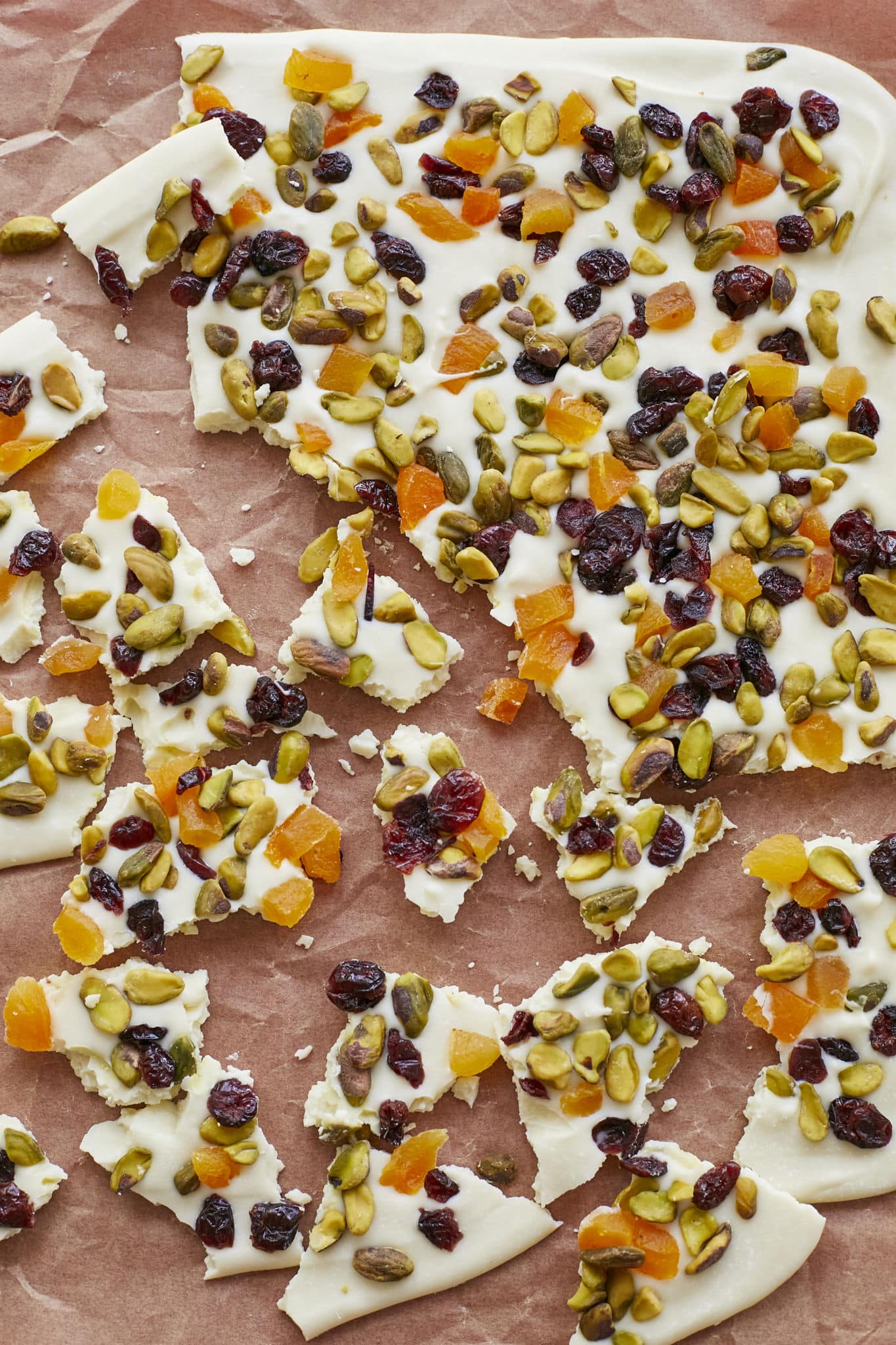 Pieces of white chocolate bark, topped with dried fruit and nuts, are broken apart on top of parchment paper. 