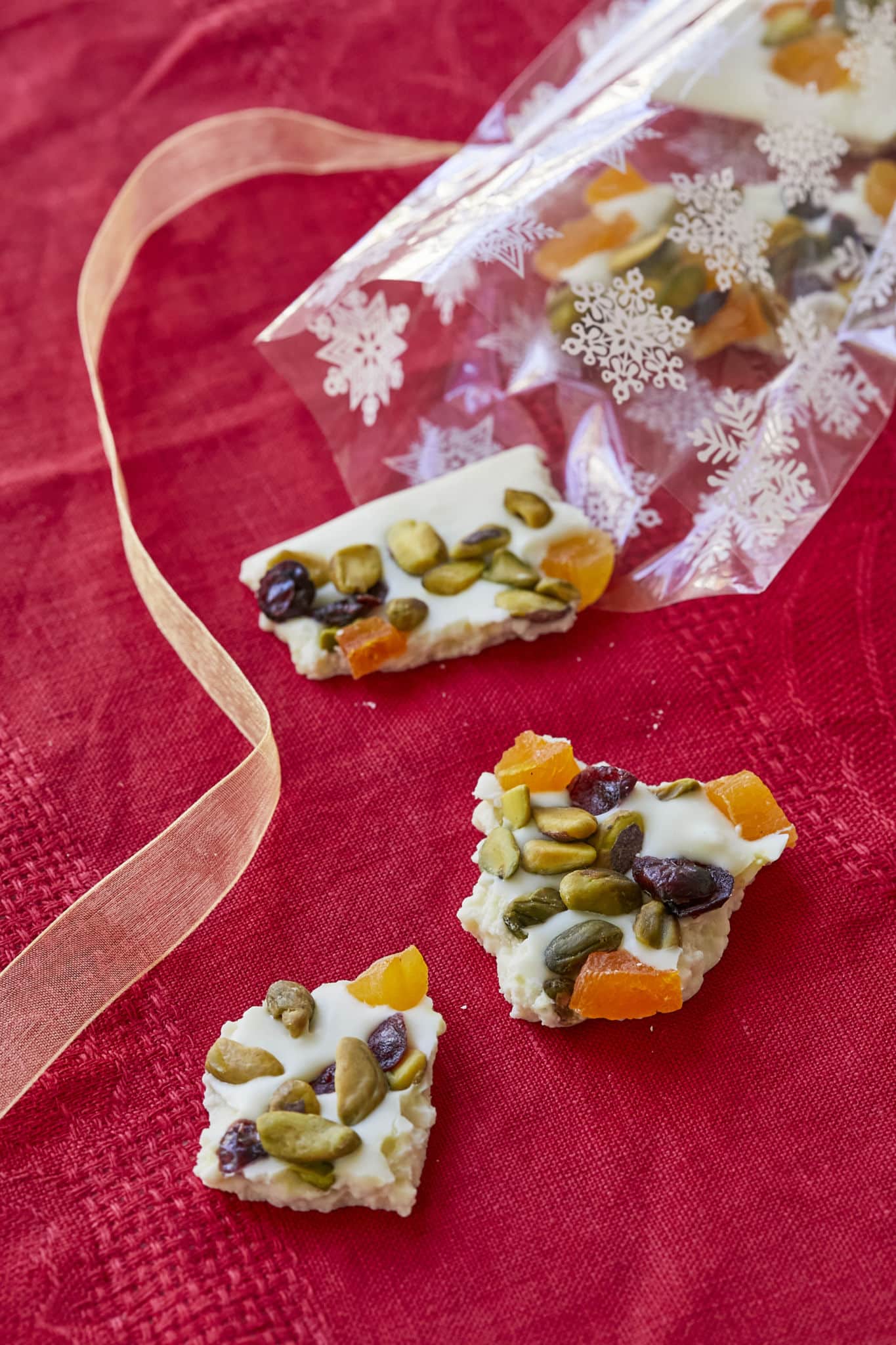 Pieces of white chocolate bark are spilled out of a clear gift bag with white snowflakes over a red table cloth.