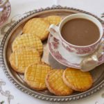 A batch of French Sable Cookies are served on a platter alongside a cup of hot tea. The butter cookies are crosshatched and golden brown.