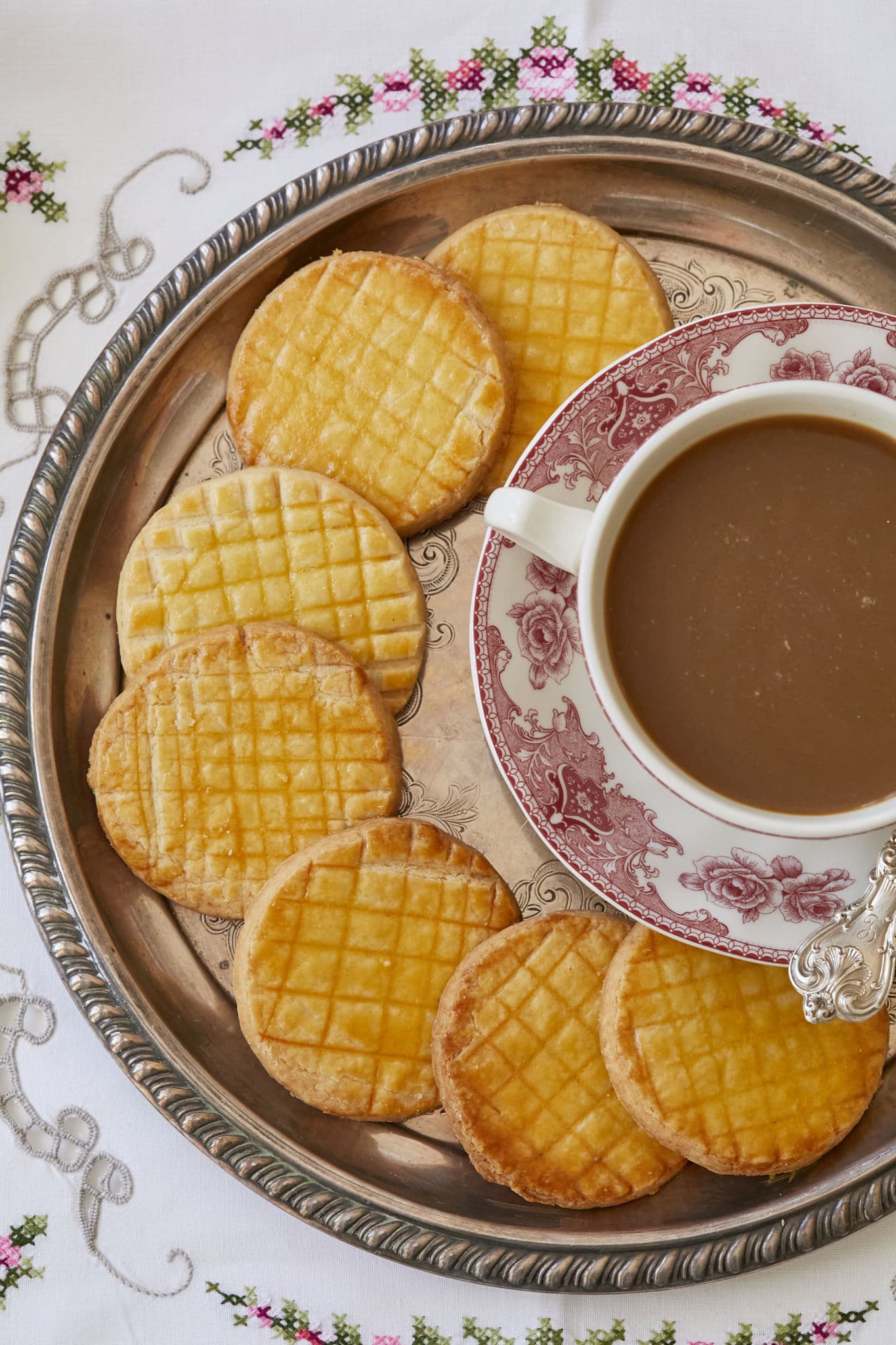 Next to a cup of tea, seven French Sable Cookies are served on a golden platter.