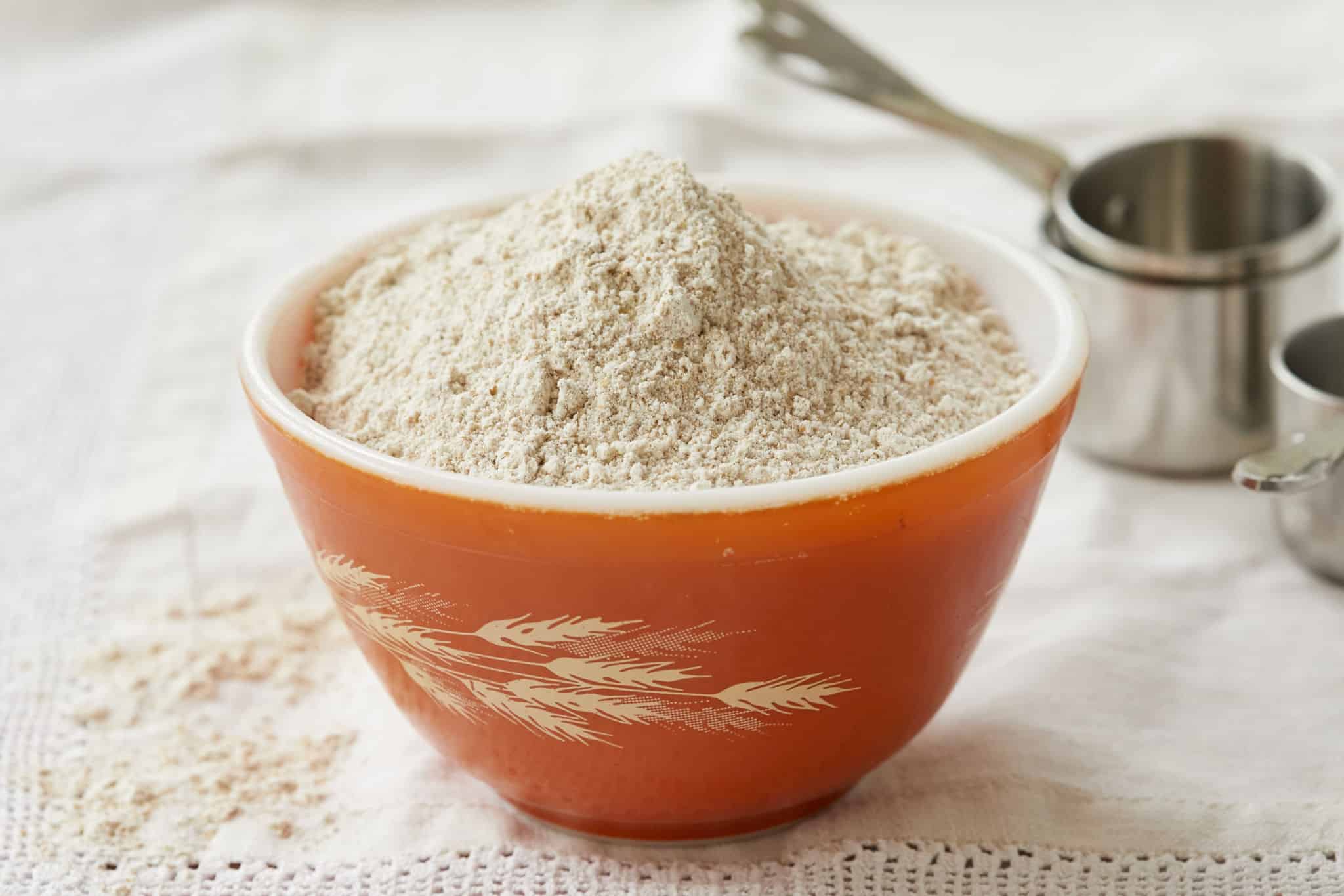 A orange Pyrex bowl with wheat grains is overflowing with whole wheat flour.