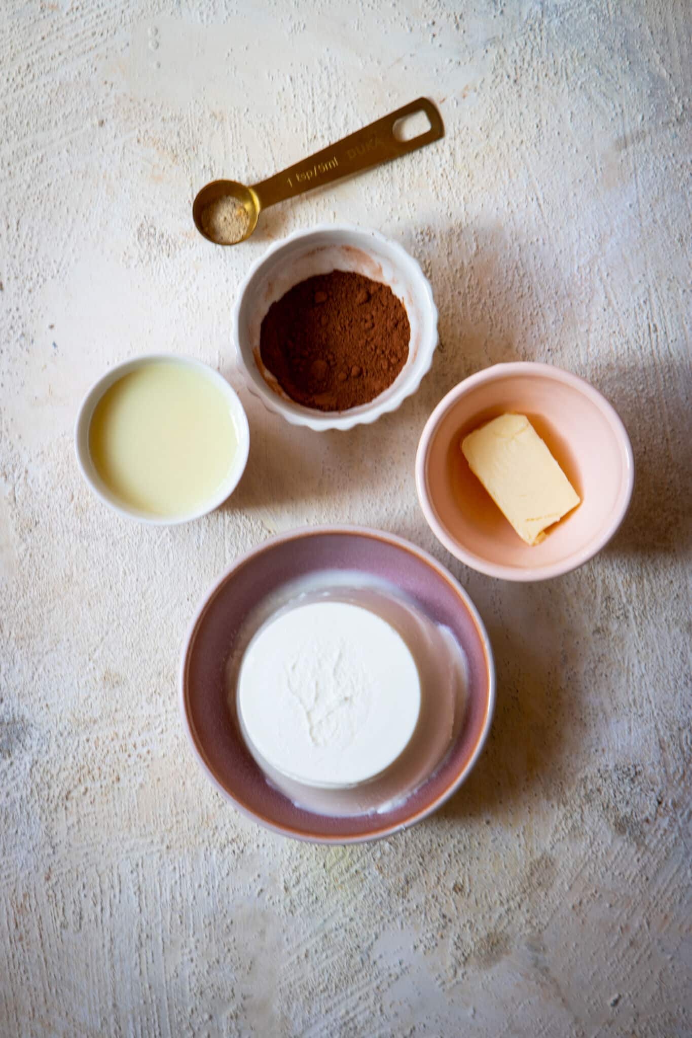 A display of the ingredients: cocoa powder, ricotta cheese, butter, and sweetened condensed milk in individual bowls with a gold teaspoon of cardamom on a table.