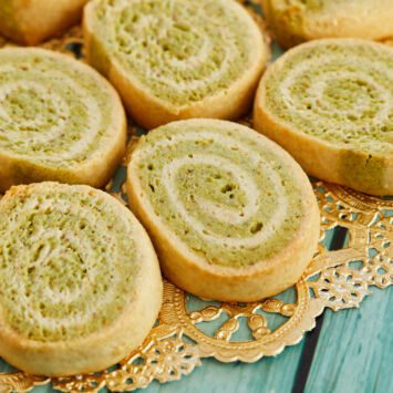 A plate of colorful kaju pista rolls on a gold doily laid out on a wooden table.