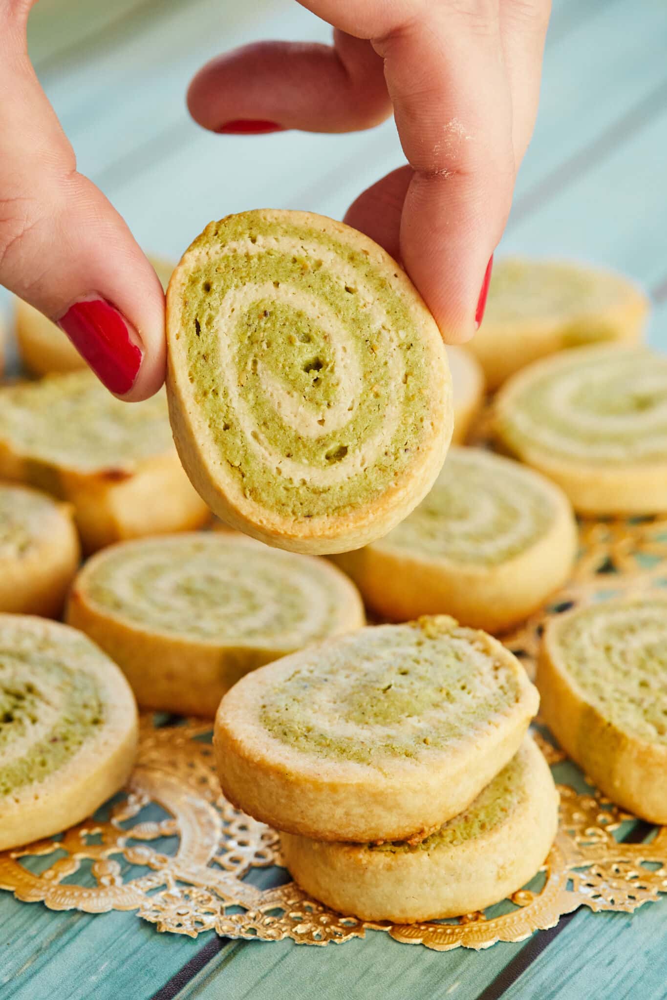 A hand holding an individual roll, with a batch of kaju pista rolls laid out on a gold doily, on a wooden table.
