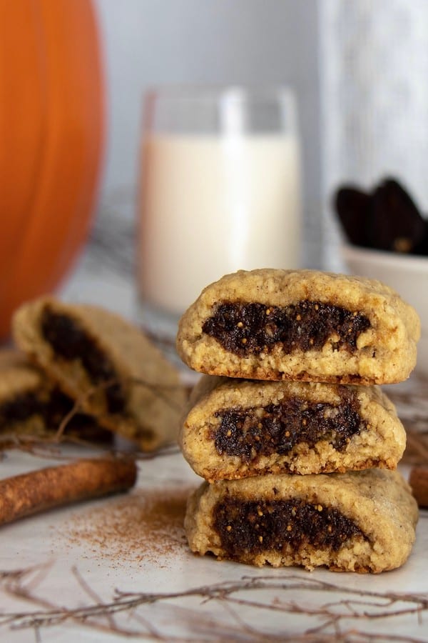 Three pumpkin spice fig bars stacked on top of each other on a counter with a glass of milk, cinnamon stick, and pumpkin in the background.