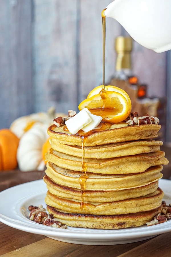 A stack of orange pumpkin pancakes with butter, chopped pecans, and orange slices, with syrup poured on top on a white plate.