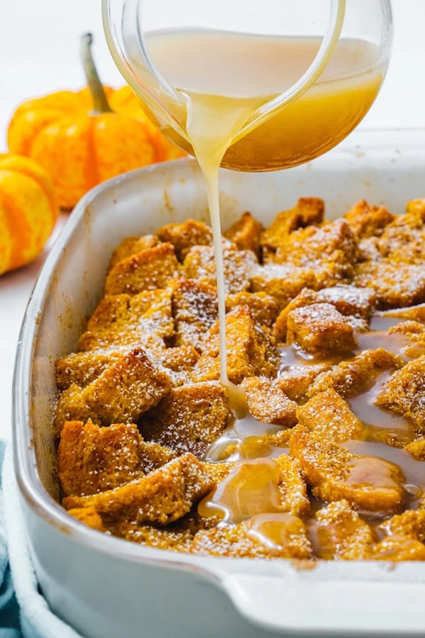 A glass dish of pumpkin bread pudding with a jug of sweet sauce being poured on top, and decorative pumpkins in the background.
