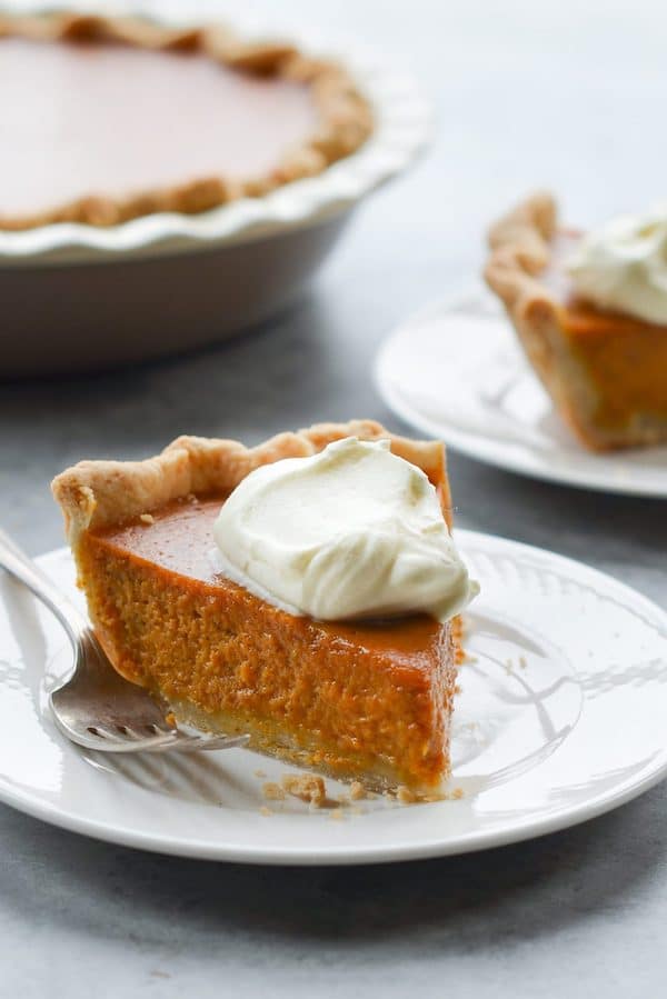 A slice of perfect pumpkin pie with a dollop of whipped cream on a white plate, in front of the pie in its pie dish.