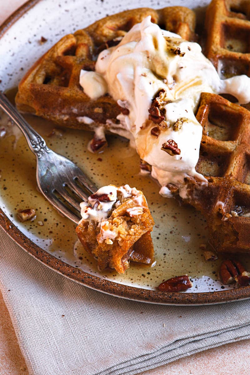 A closeup image of a partially eaten pumpkin waffle. The waffle is topped with whipped cream, maple syrup, and toasted pecans.