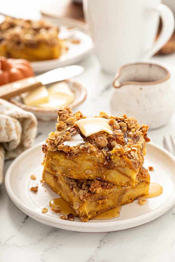 Two pieces of French toast casserole stacked on top of each other on a white plate, with melted butter and syrup on top of a fall-inspired table.