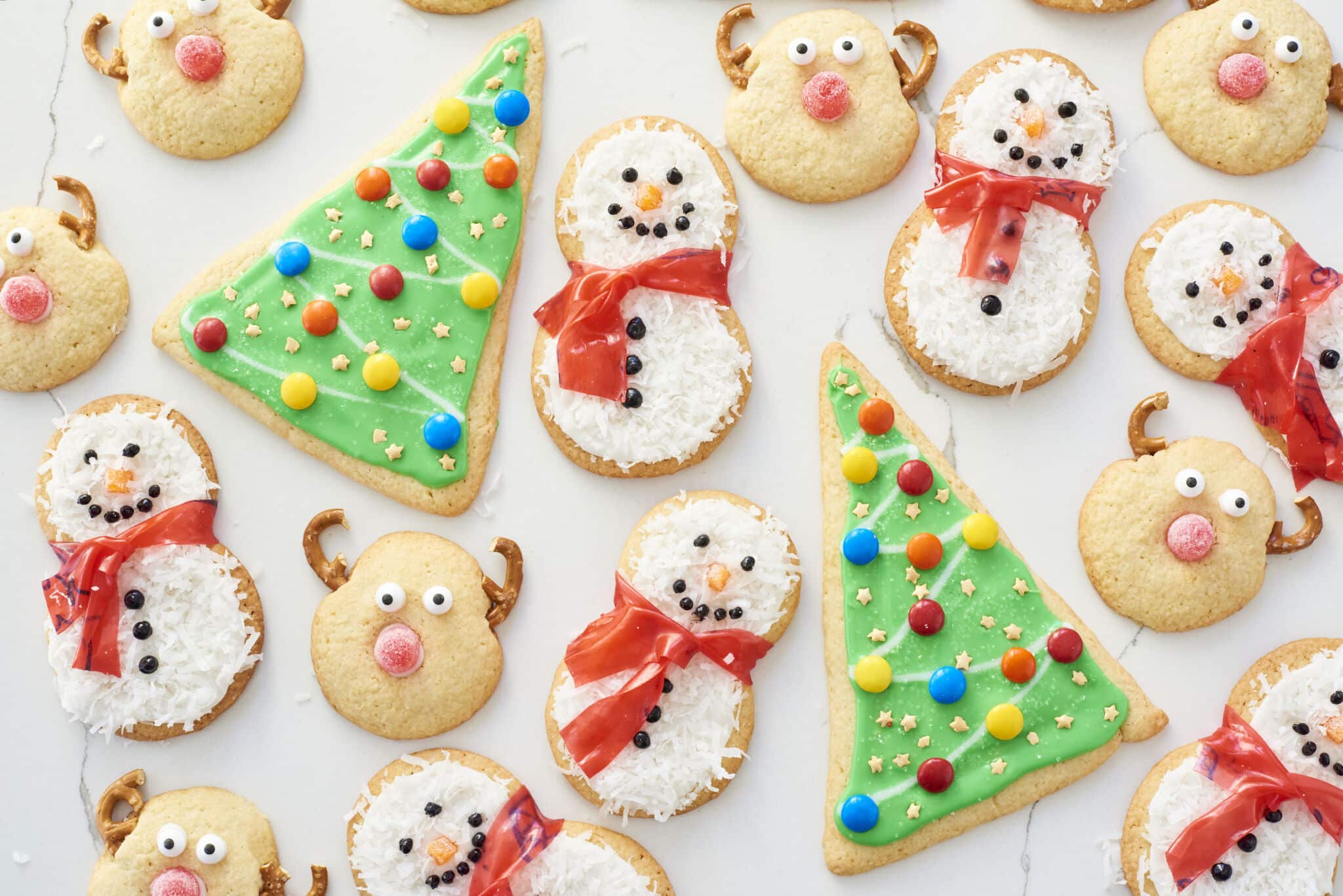 Three differently decorated sugar cookies are presented on a white table. They are Christmas Tree Cookies, Snowman Cookies, and Reindeer Cookies.
