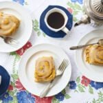 Three homemade Pumpkin Cinnamon Buns are served on white dishes with mugs of black coffee.