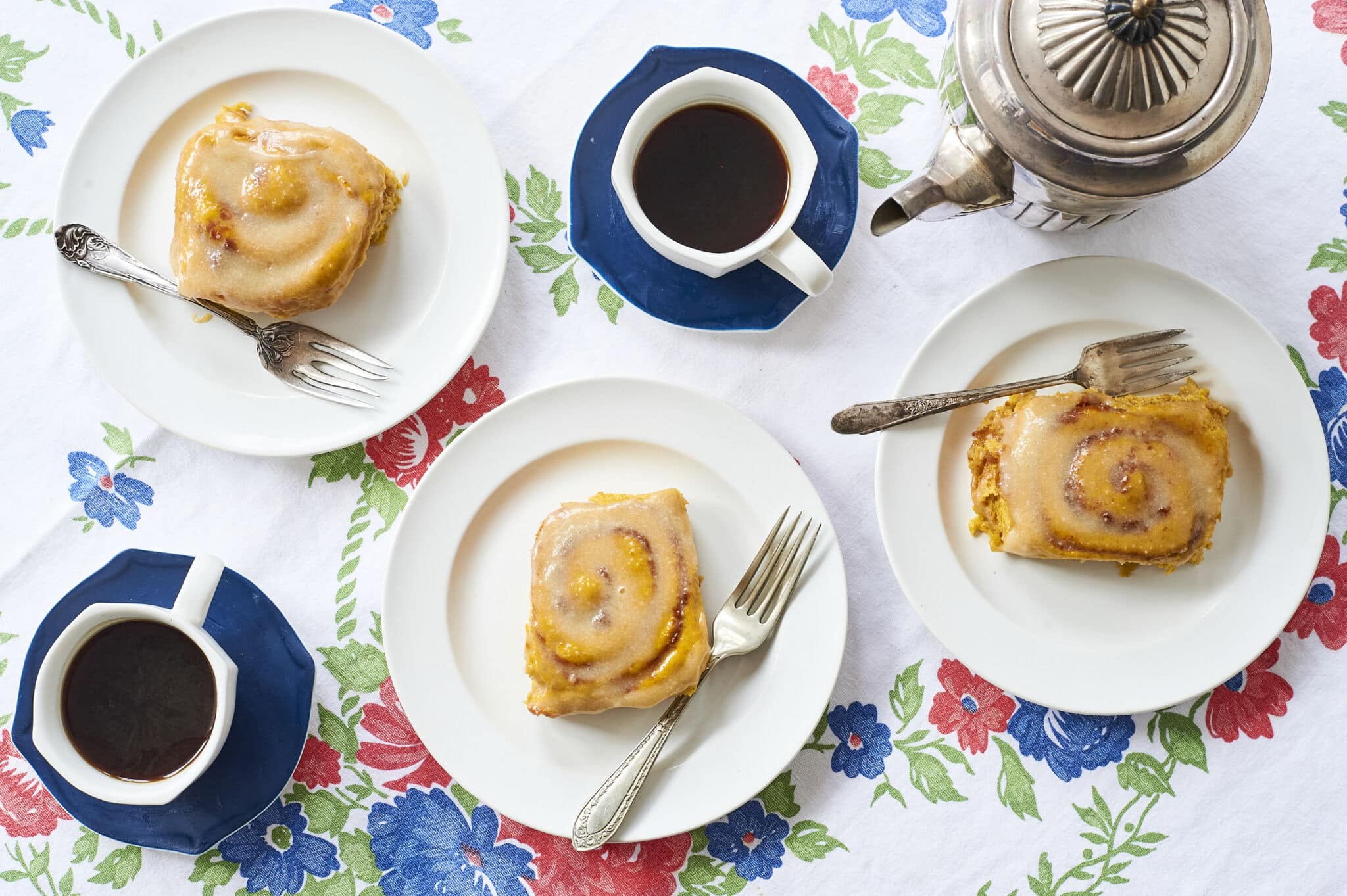 Three homemade Pumpkin Cinnamon Buns are served on white dishes with mugs of black coffee.