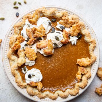 Chai pumpkin pie from overhead, with a scalloped, flaky crust and topped with crust leaf designs, whipped cream, and pumpkin seeds, nest to a small glass bowl of cinnamon sugar on a white table.