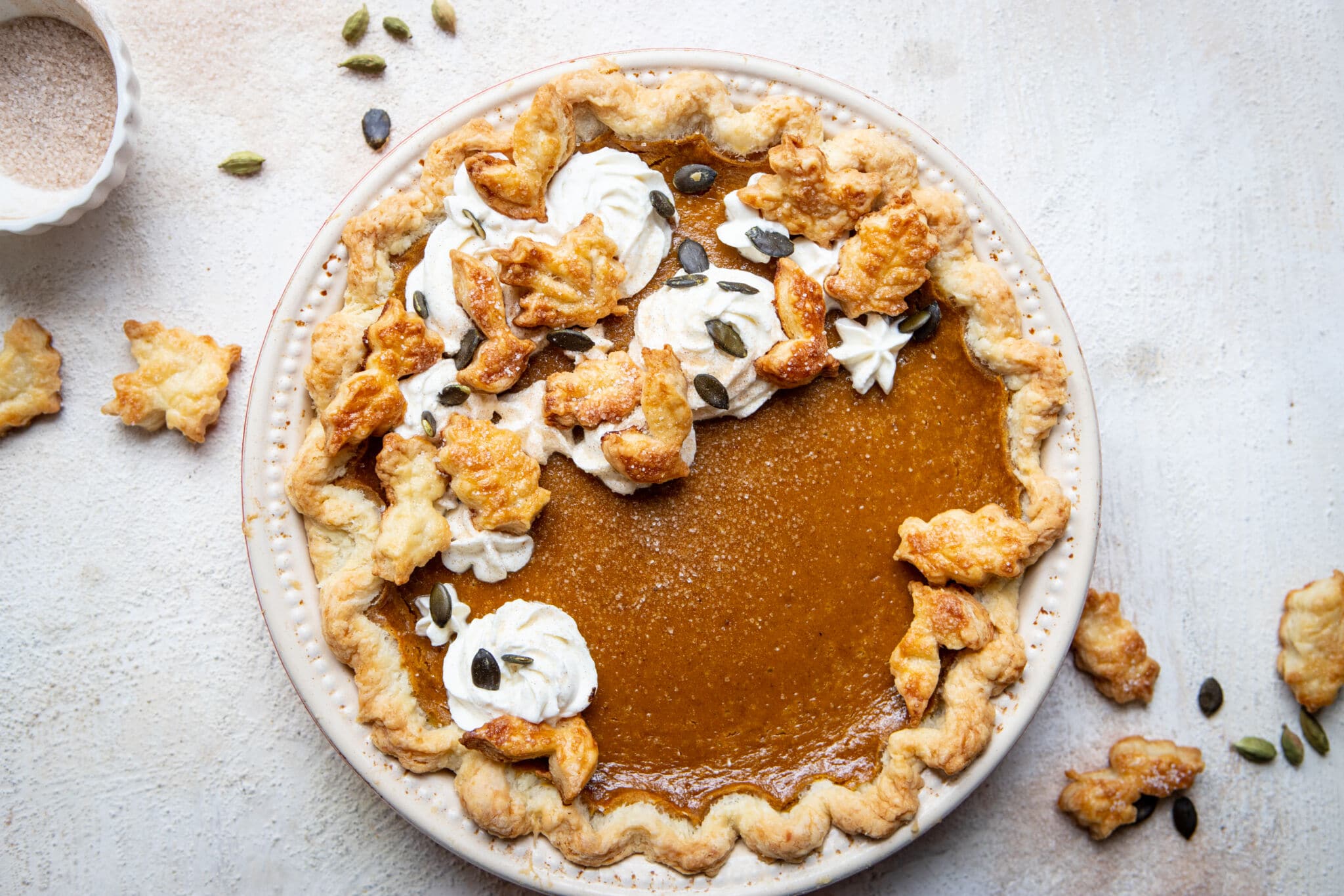 Chai pumpkin pie from overhead, with a scalloped, flaky crust and topped with crust leaf designs, whipped cream, and pumpkin seeds, nest to a small glass bowl of cinnamon sugar on a white table.