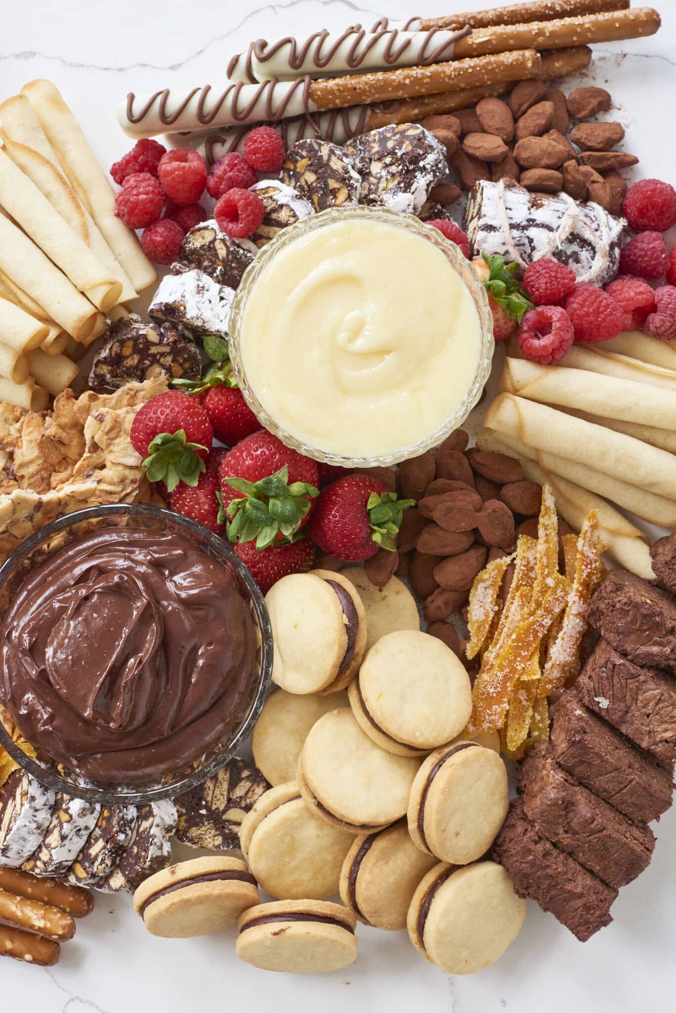 A top down view of the arranged dessert charcuterie board with chocolate dips, cookies, fresh fruit, and nuts on a marble serving tray.