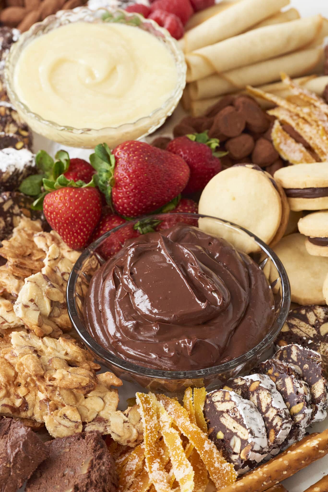 A close up of elements of the dessert charcuterie board including white chocolate ganache dip, chocolate ganache dip, strawberries, sandwich cookies, almond brittle, chocolate salami, and candied fruit.
