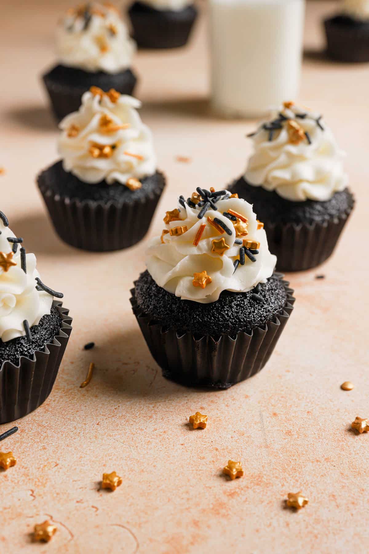 Deep, dark chocolate cupcakes are served on a table top in front of a glass of milk. They are decorated with white frosting and festive black and gold sprinkles.