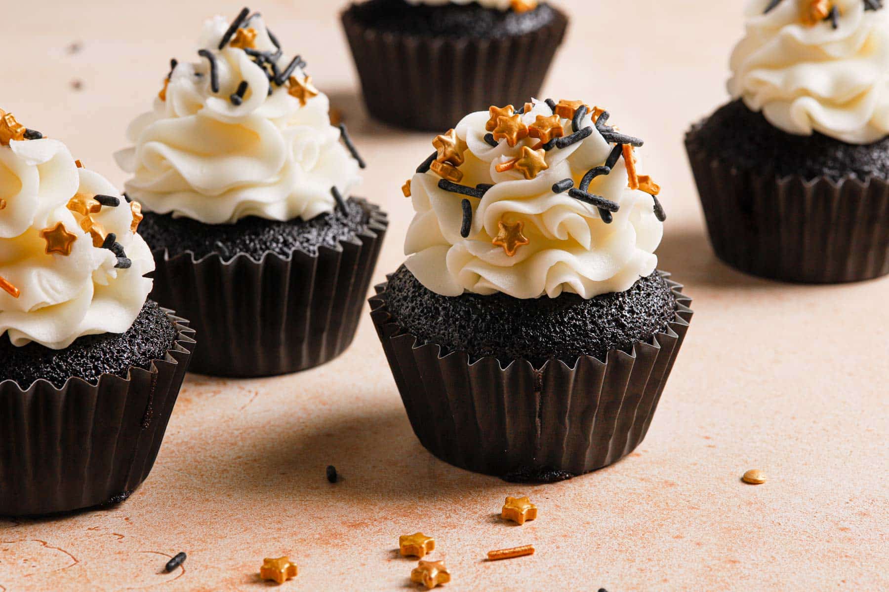 Homemade Black Cocoa Cupcakes are decorated with white meringue cream and topped with black, gold, and gold star sprinkles.